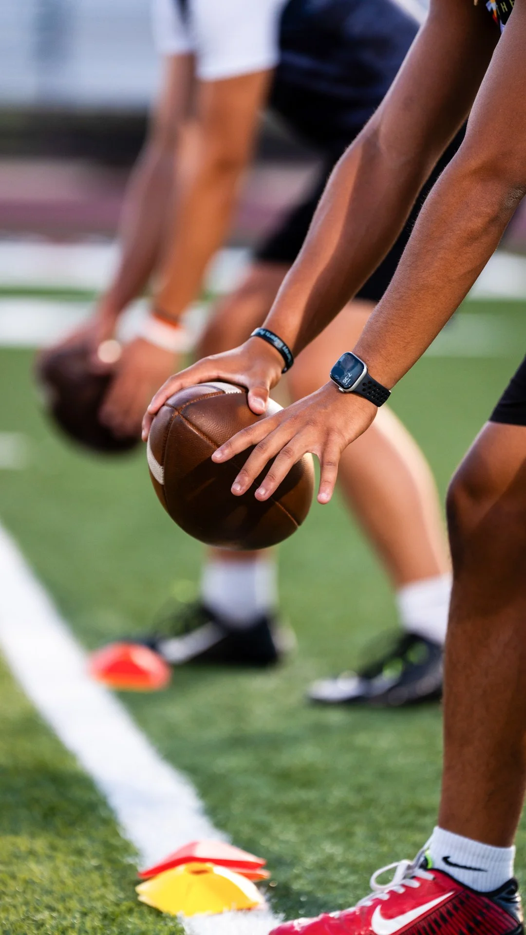 Football players in a stance, preparing for a snap during practice or game, with cones on the field.