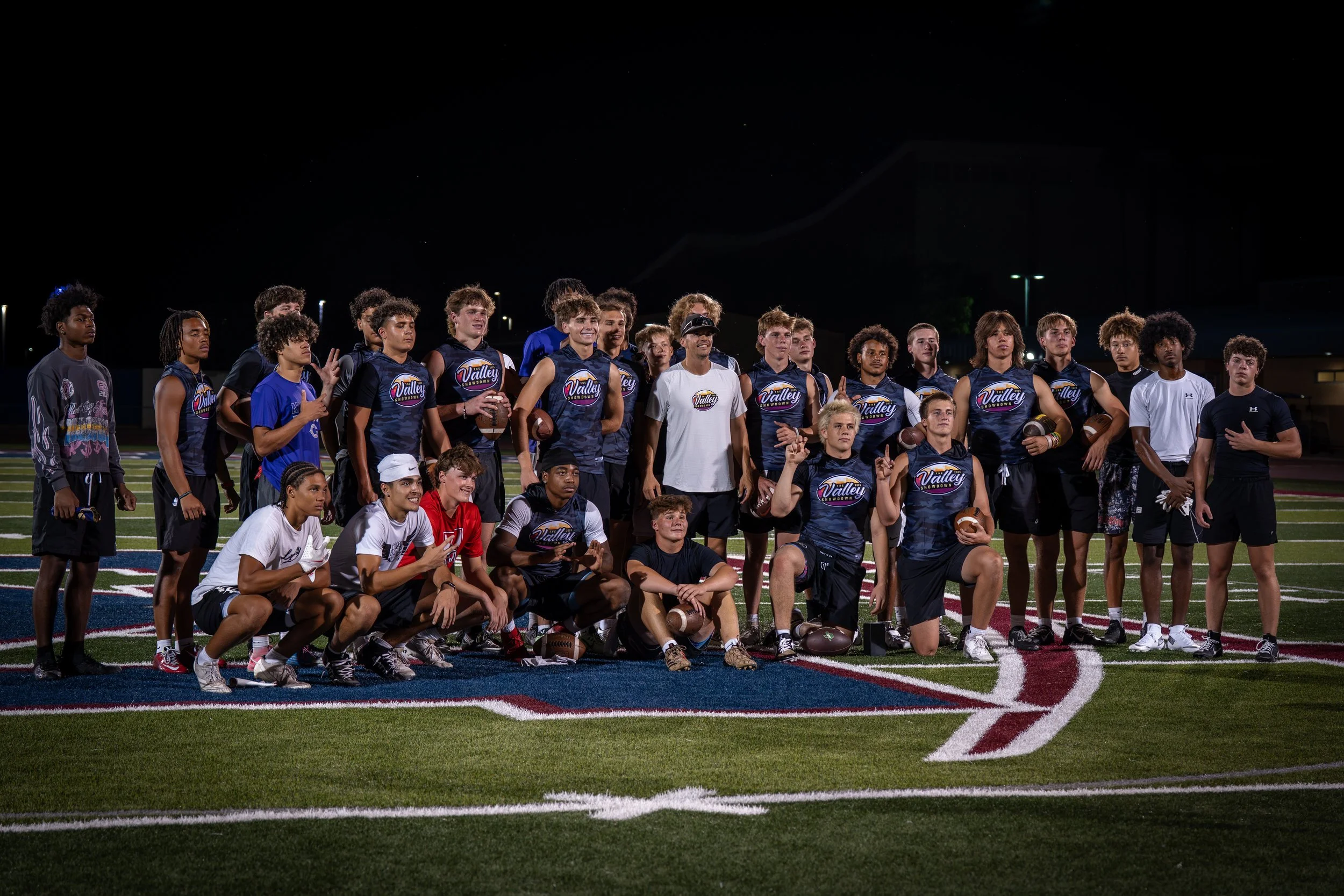 A group of young football players and coaches posing on a sports field at night, some holding footballs, wearing team uniforms.
