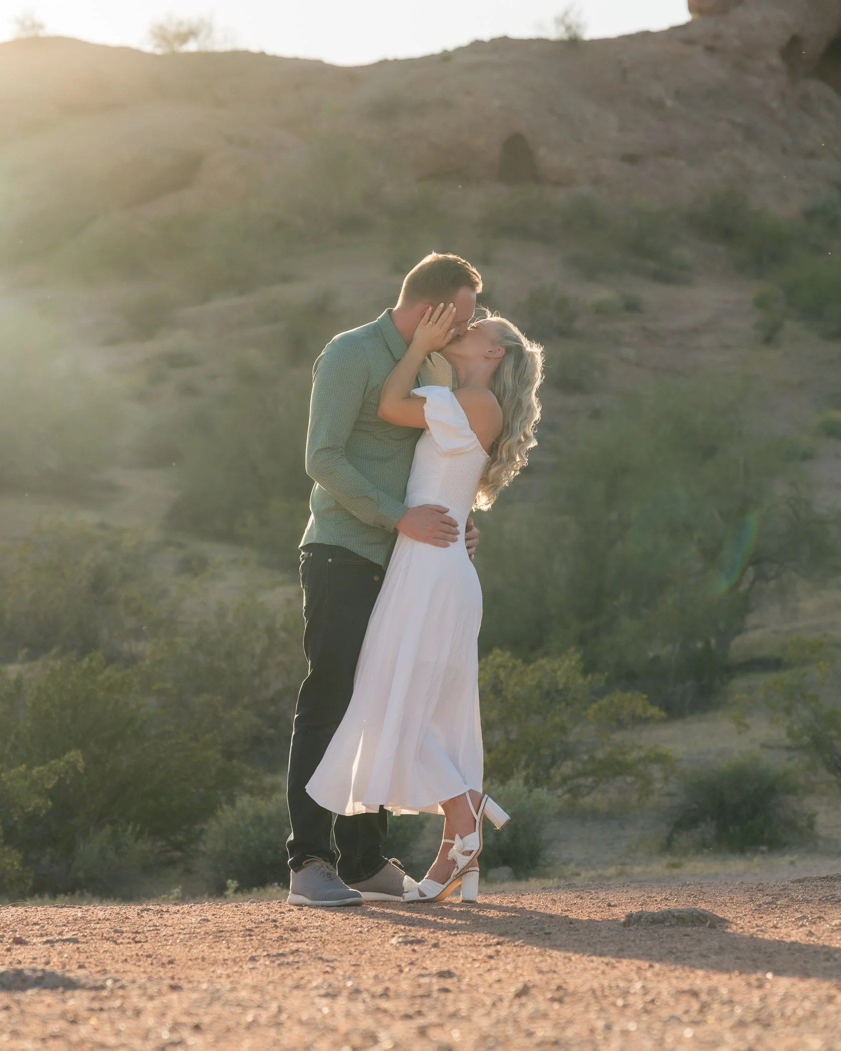 A couple sharing a kiss outdoors on a dirt path with a hilly landscape in the background, during sunset.