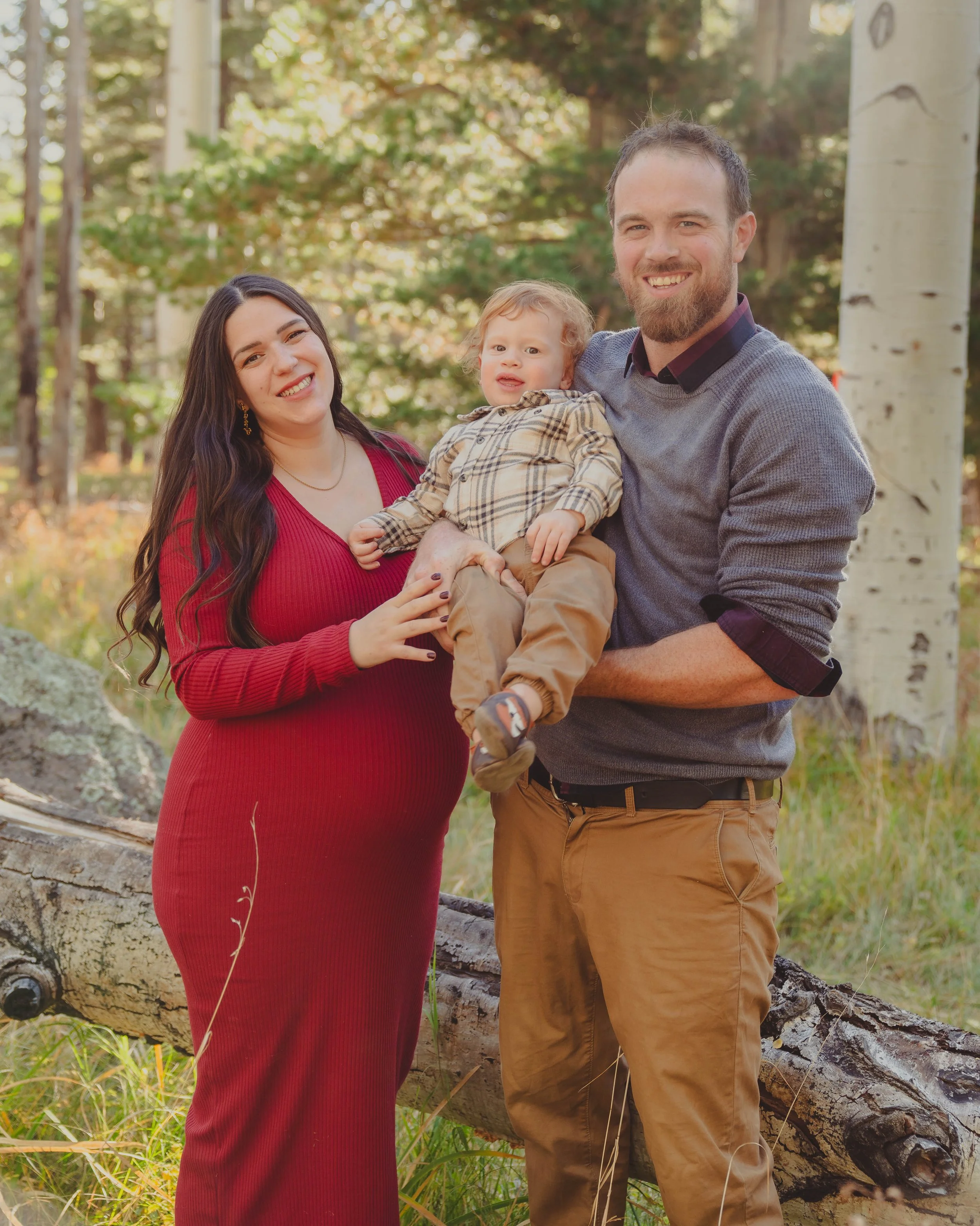 A family of three, a woman in a long red dress, a man in a gray sweater and khakis, and a toddler in a plaid shirt, standing outdoors in a forested area with trees and greenery.