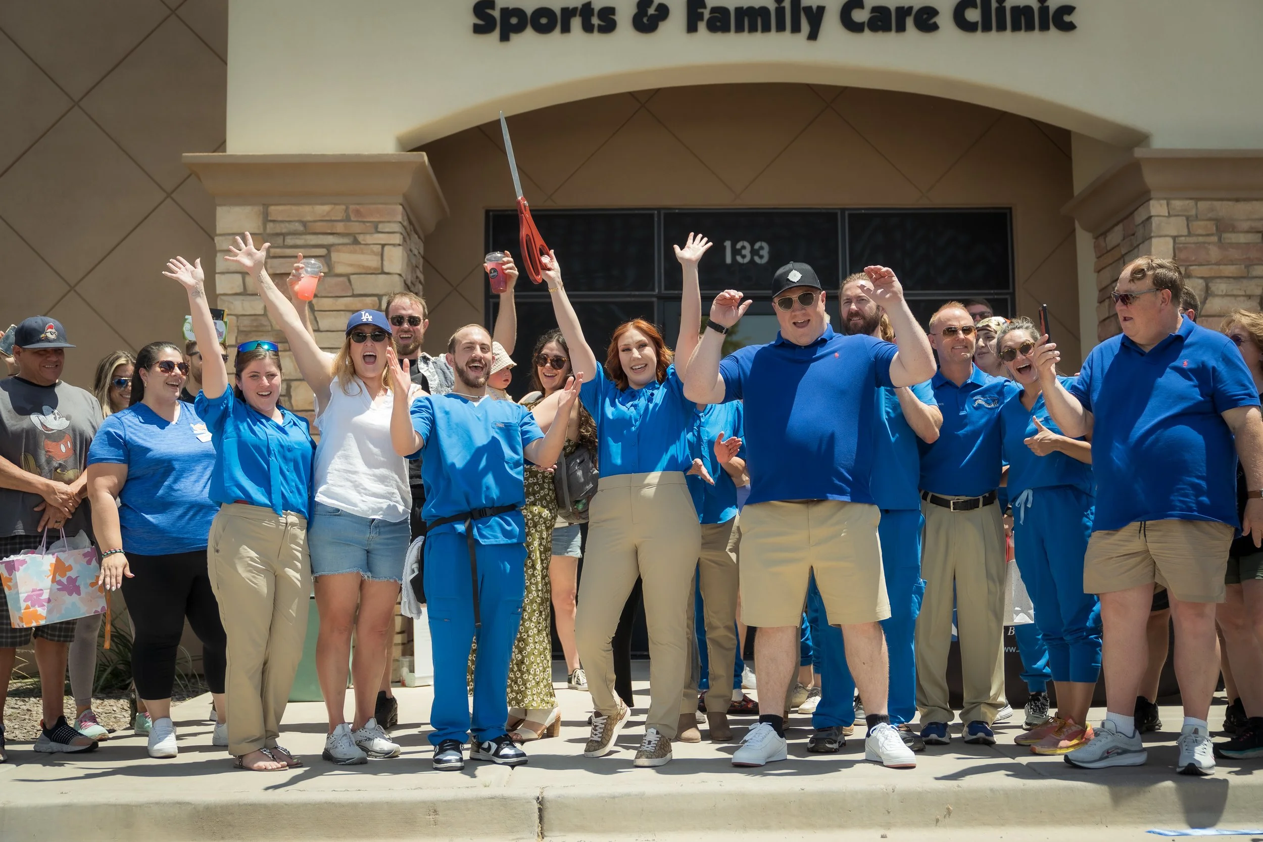 Group of people standing outside a building with a sign that reads 'Sports & Family Care Clinic', celebrating and smiling, some waving, with some dressed in blue uniforms, one holding scissors and another with drinks.