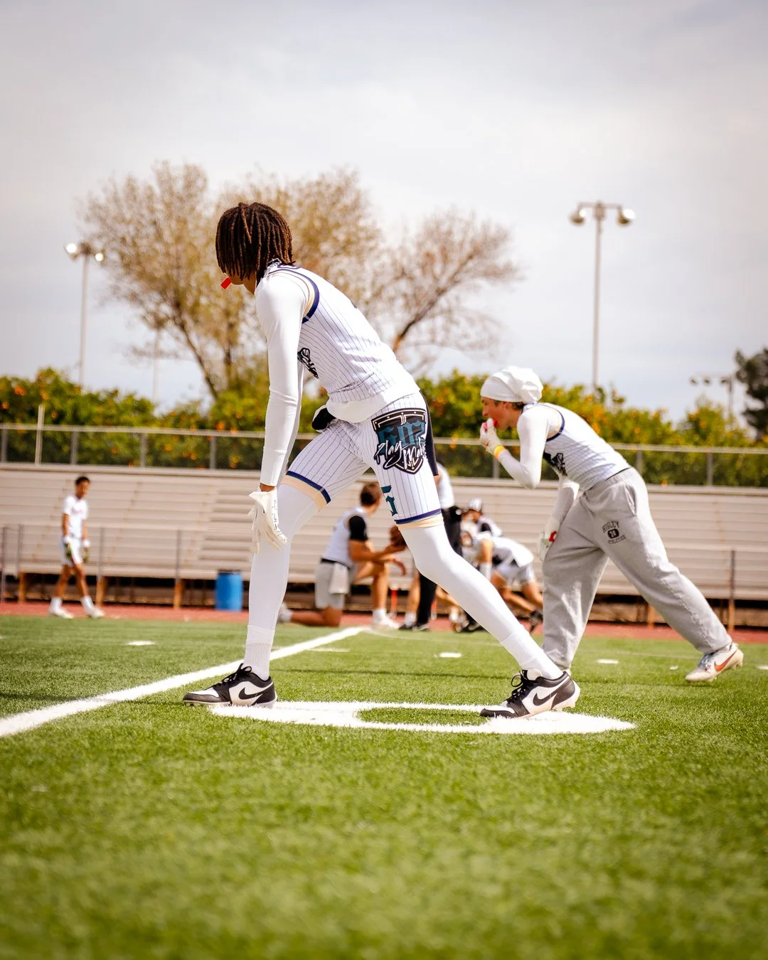Two football players in white uniforms preparing to start a game on a sports field with other players and coaches in the background.