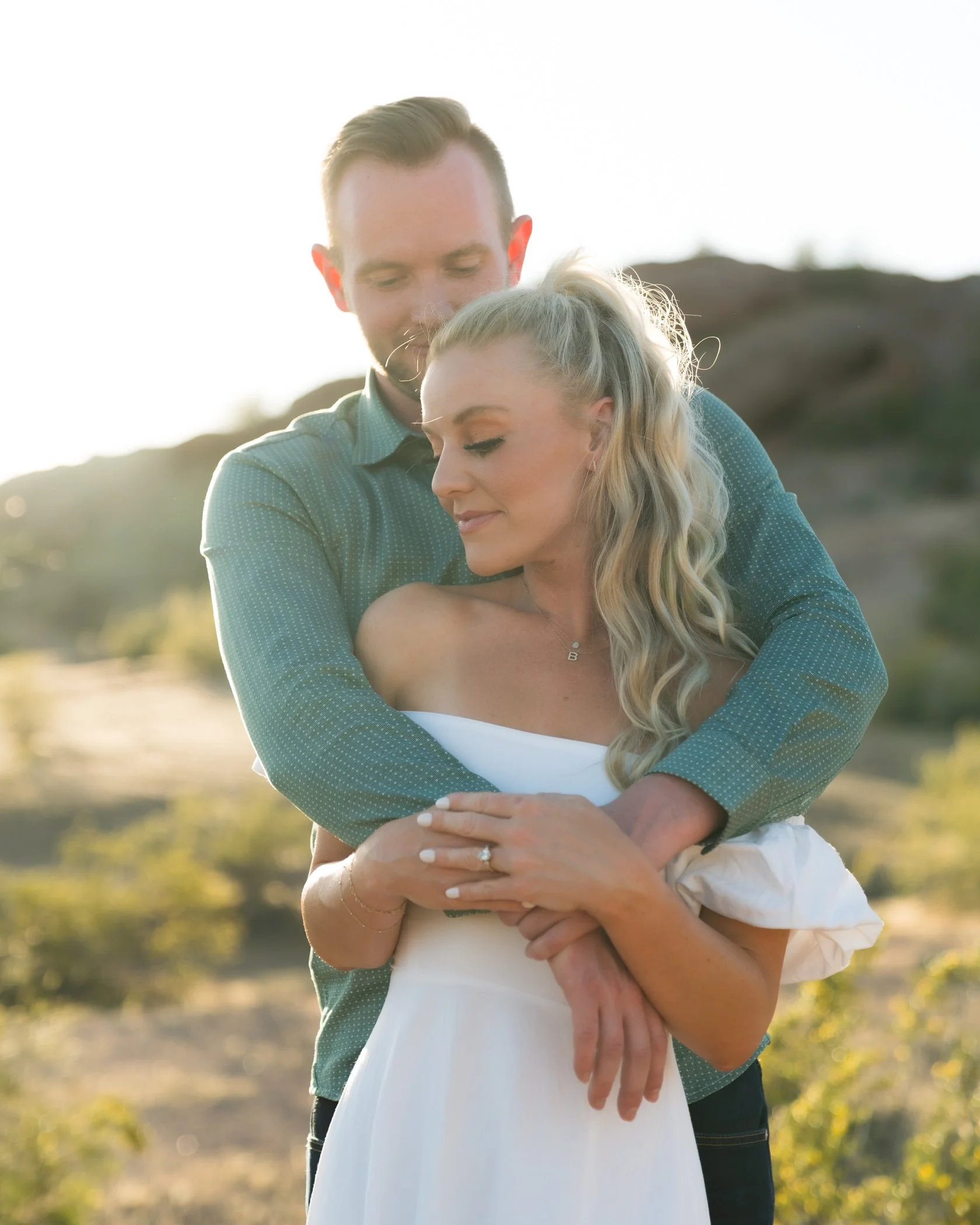 A couple embracing outdoors during sunset, the man with his arms around the woman who is smiling with her eyes closed, in a scenic natural setting.