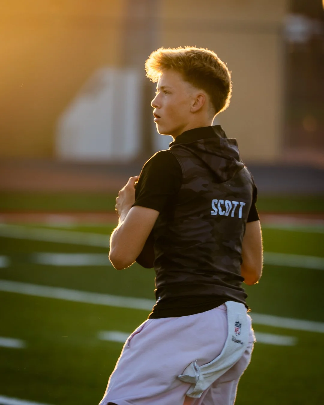 A young man wearing a black shirt with the word 'SSDT' on the back, white shorts, standing on a sports field during sunset, holding his shoulder, and looking to the side.