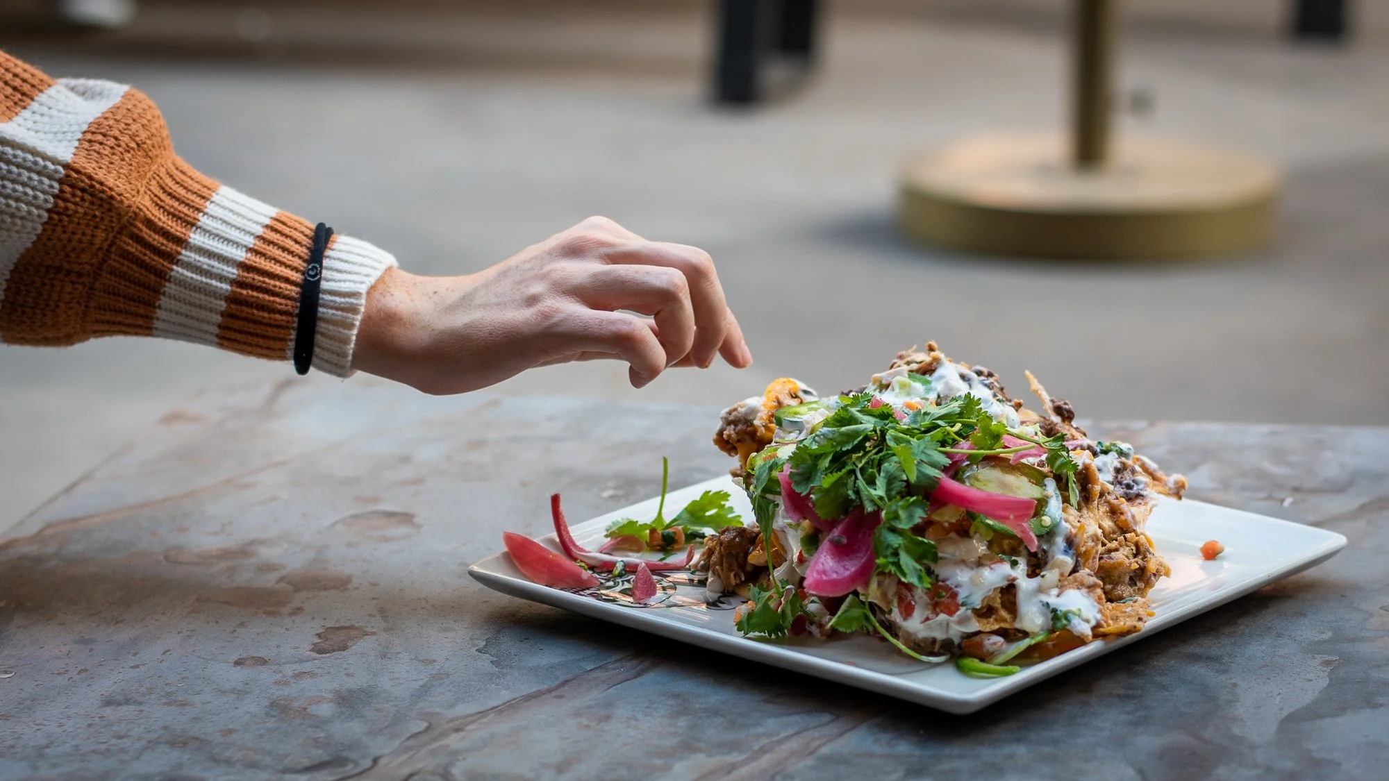 Person's hand reaching to touch a large serving of Mexican food garnished with cilantro, radish slices, and sauce on a white plate outdoors.
