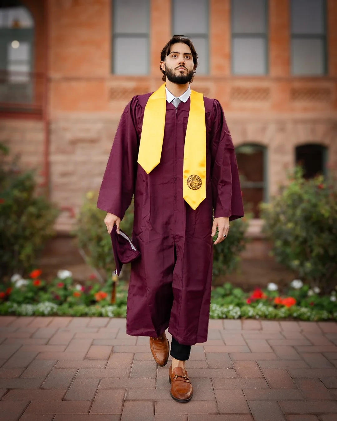 A young man in a burgundy graduation gown and yellow stole standing outdoors in front of a brick building and flowerbeds.
