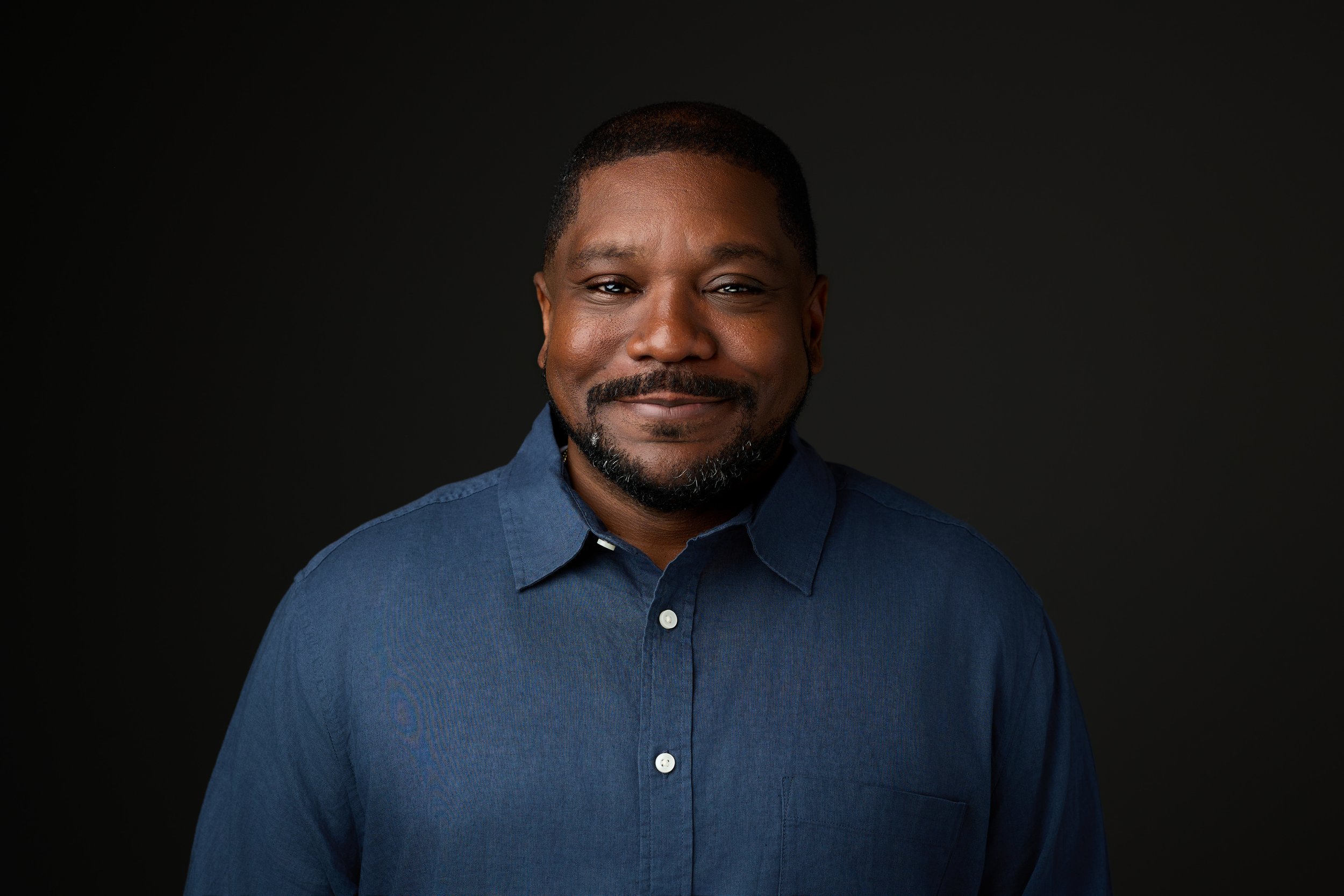 A middle-aged African American man with short hair and a goatee, wearing a dark blue button-down shirt, smiling against a dark background.