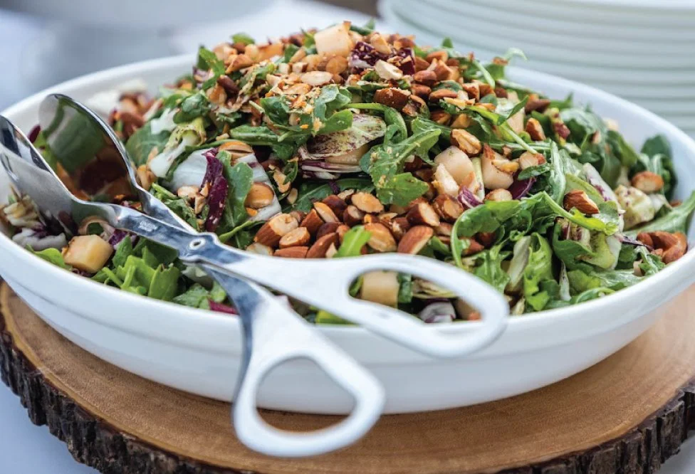 A fresh mixed salad with greens, chopped vegetables, and chopped nuts in a white bowl with metal tongs resting on the edge.