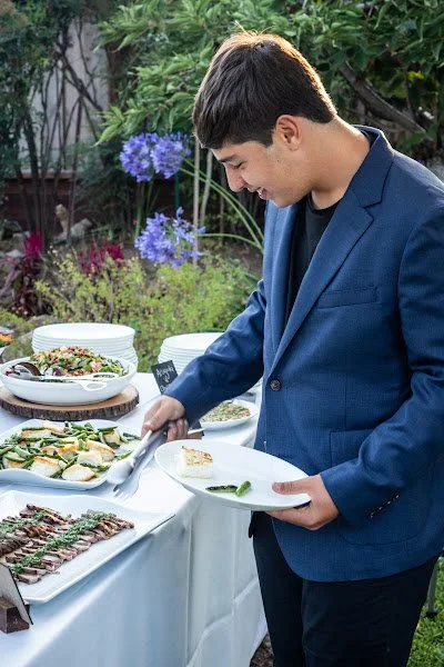 A bar mitzvah boy in a blue blazer serves himself food at a buffet outdoors, with dishes of salads and grilled meat on the table, surrounded by greenery and purple flowers.