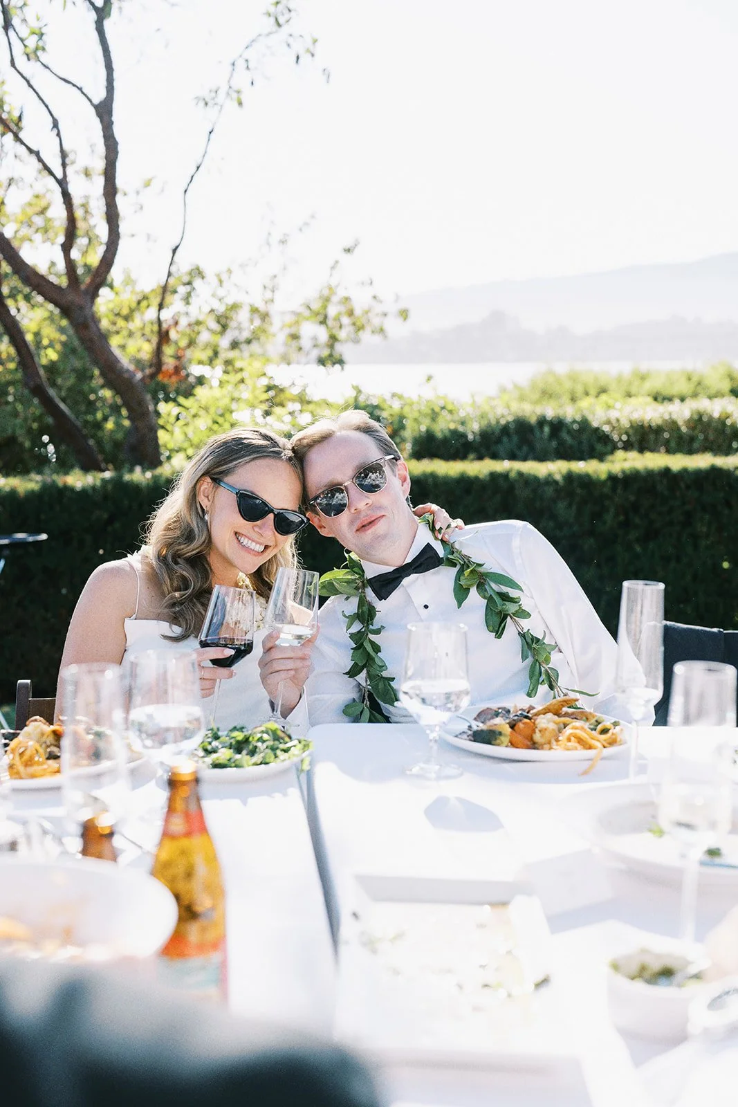 Bride and  groom enjoying an outdoor wedding, holding glasses of wine, with food on the table and lush greenery in the background.