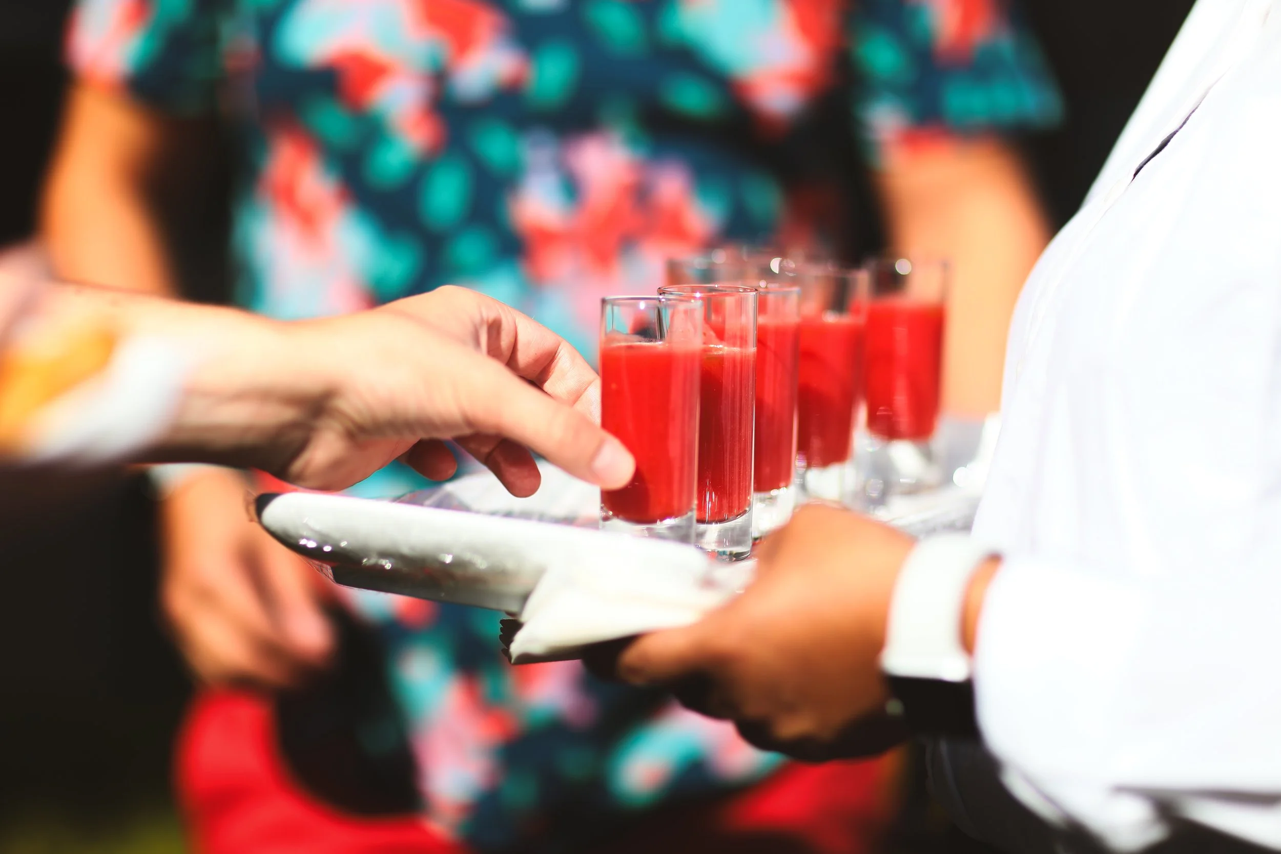 A person holding a tray with multiple glasses filled with gazpacho.
