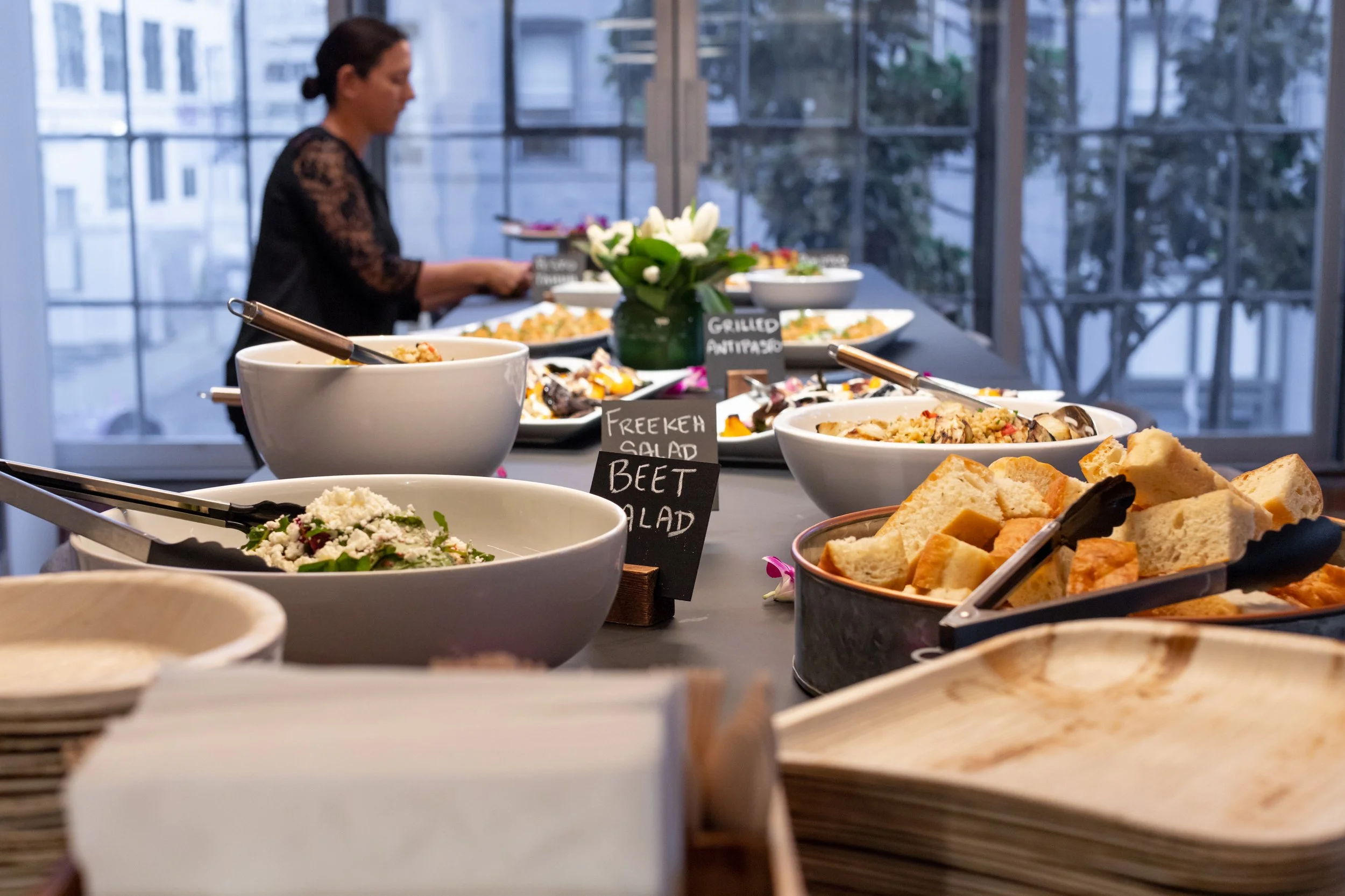 A buffet table with various salads, breads, and dishes set up in a bright room with large windows, with a woman in the background serving herself.