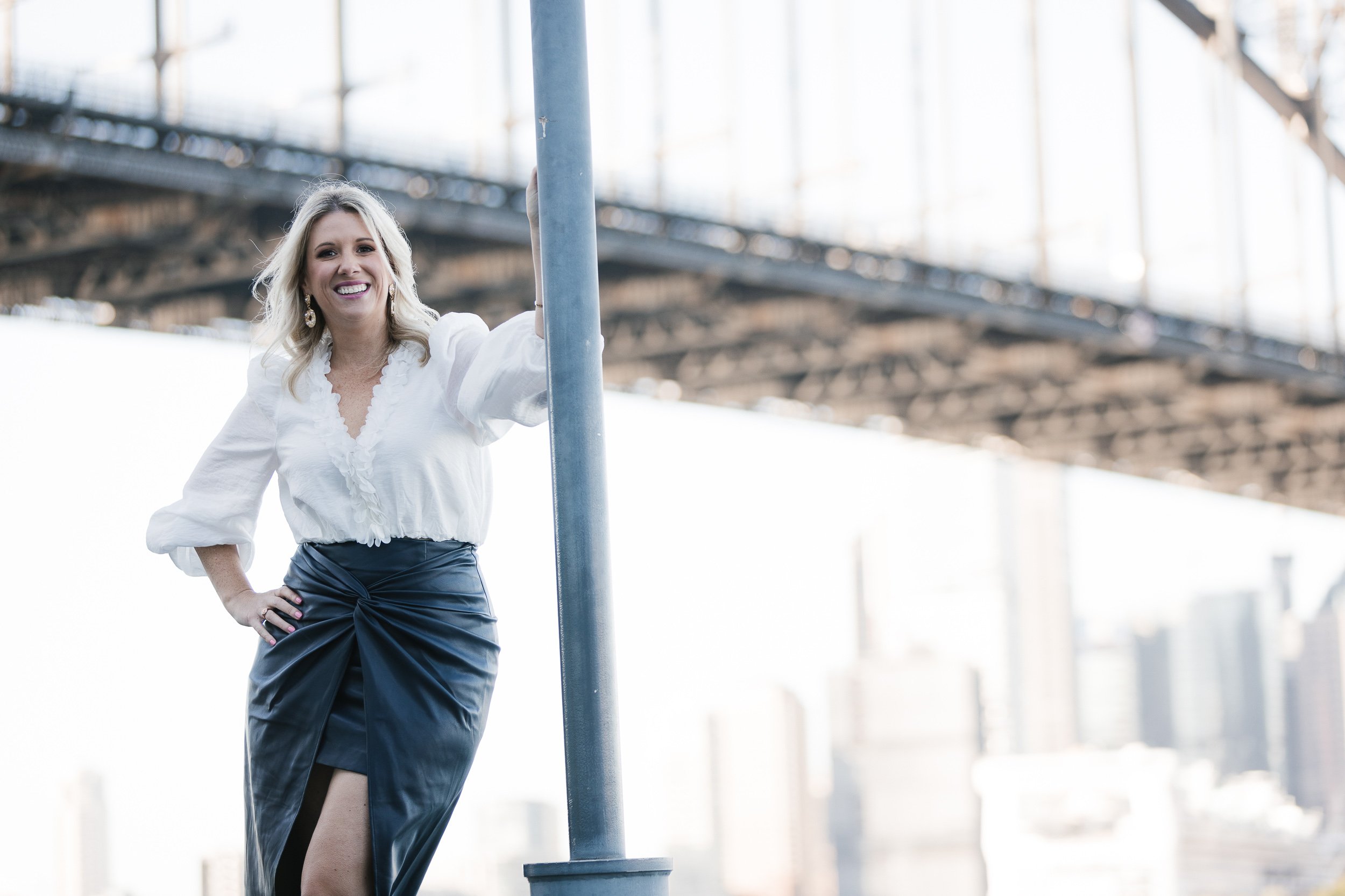 A woman with blonde hair wearing a white blouse and a black leather skirt, standing under a bridge in an urban setting, smiling at the camera.