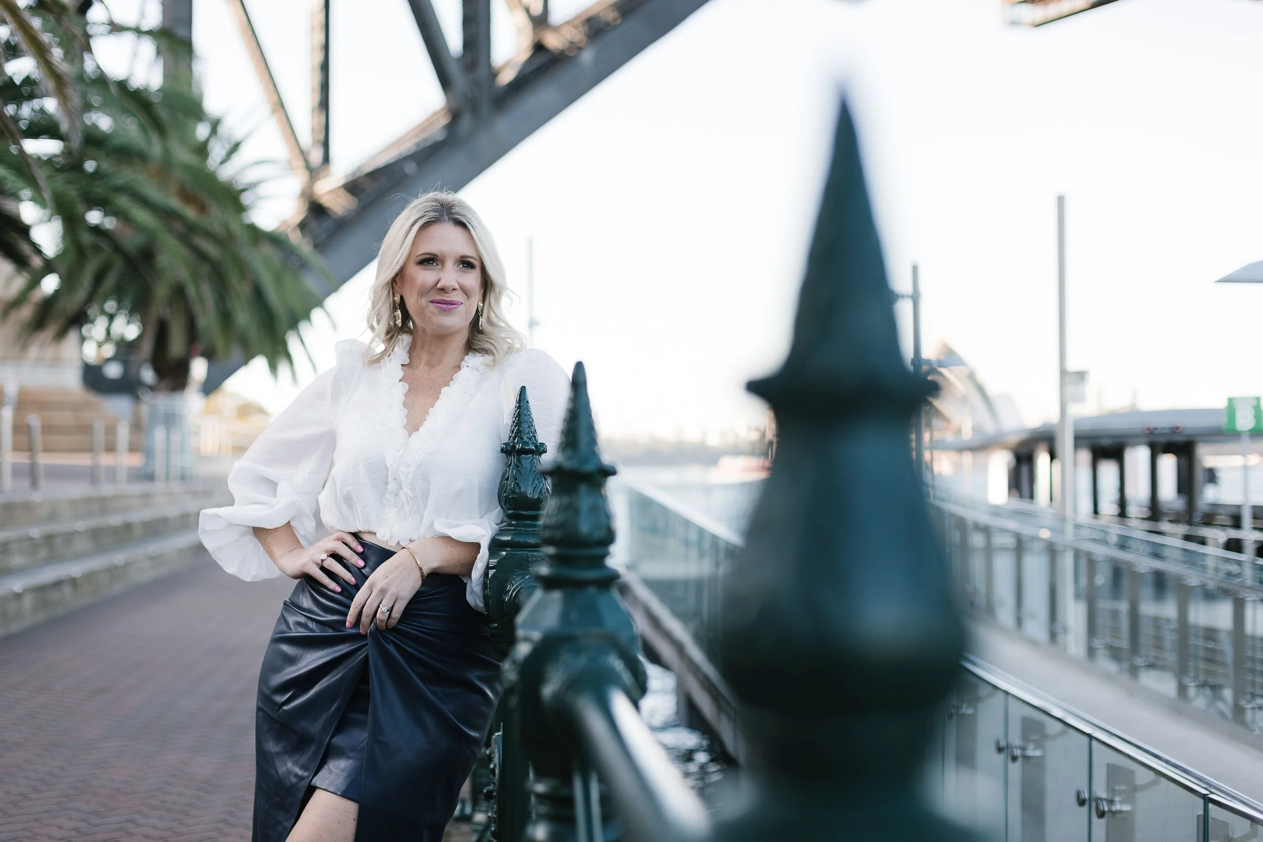 A woman with blonde hair wearing a white blouse and black skirt leaning against a railing near a waterfront with bridges and boats in the background.