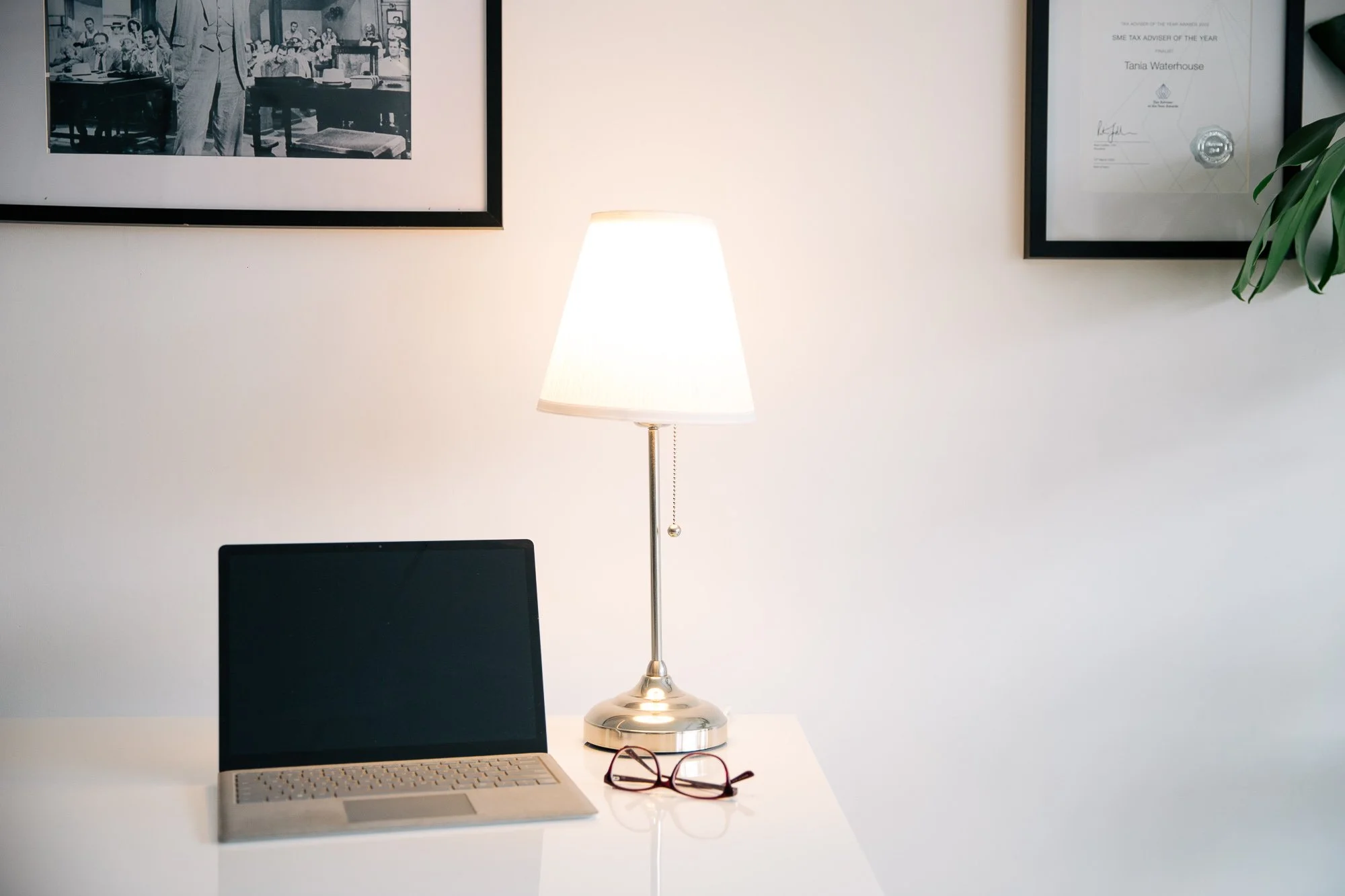 A laptop, a pair of glasses, a lamp, and framed pictures on a white desk and wall.