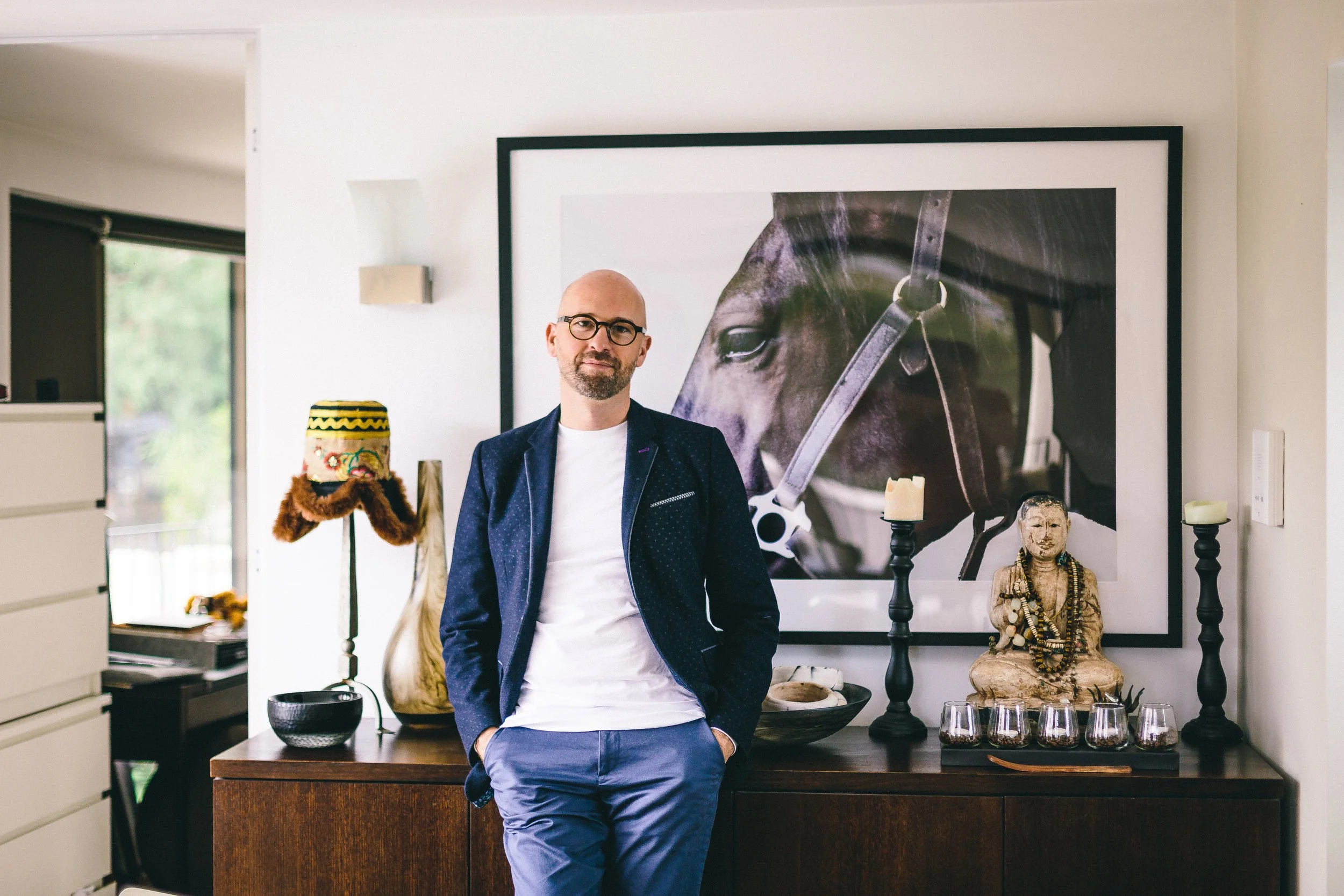 A man in a navy blazer and blue pants stands in front of a wooden sideboard in a home interior with eclectic decor, including a large horse portrait, candles, a Buddha statue, and decorative objects.