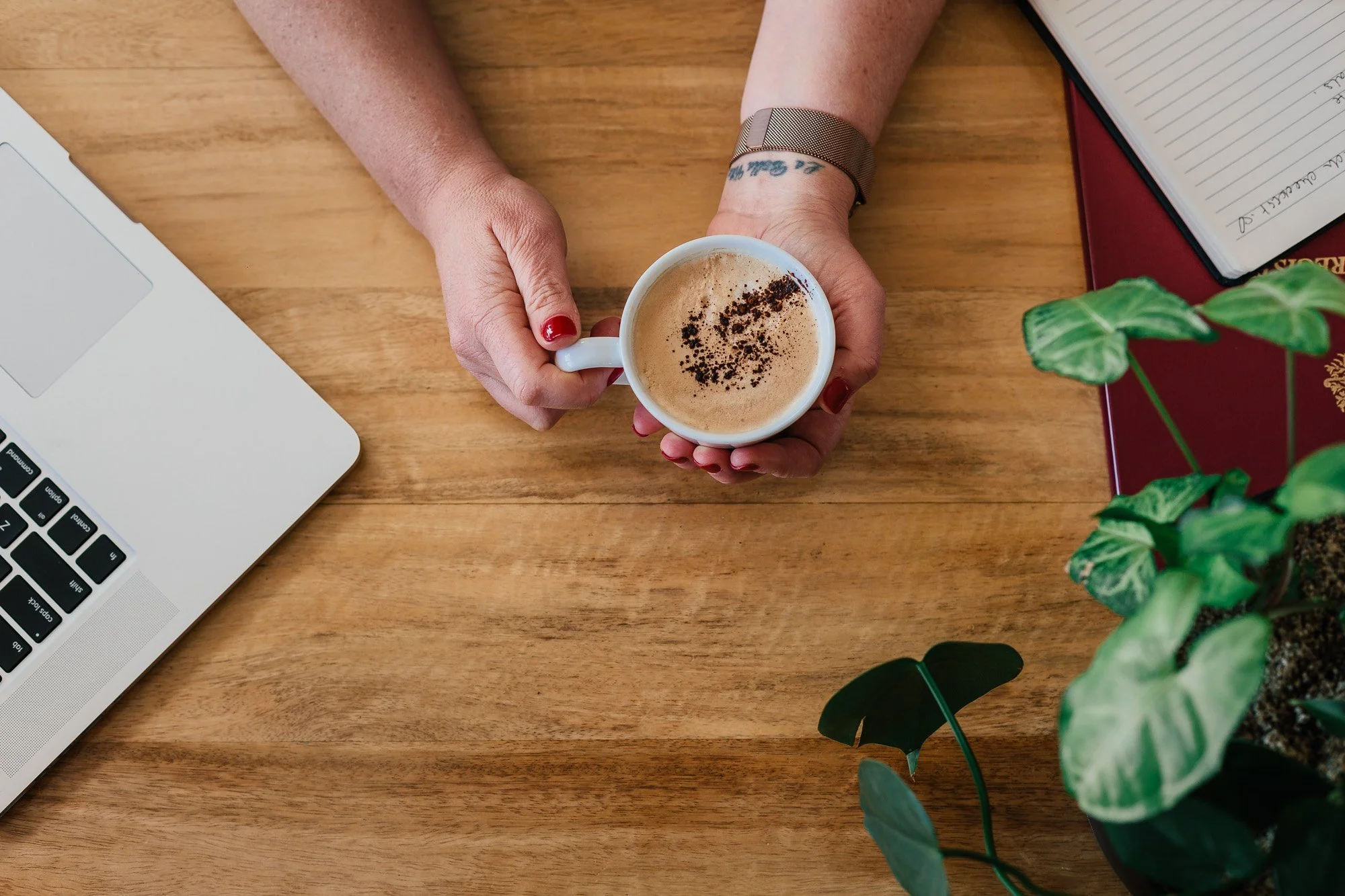 A person holding a cup of coffee on a wooden table, with a laptop, notebooks, and a plant nearby.