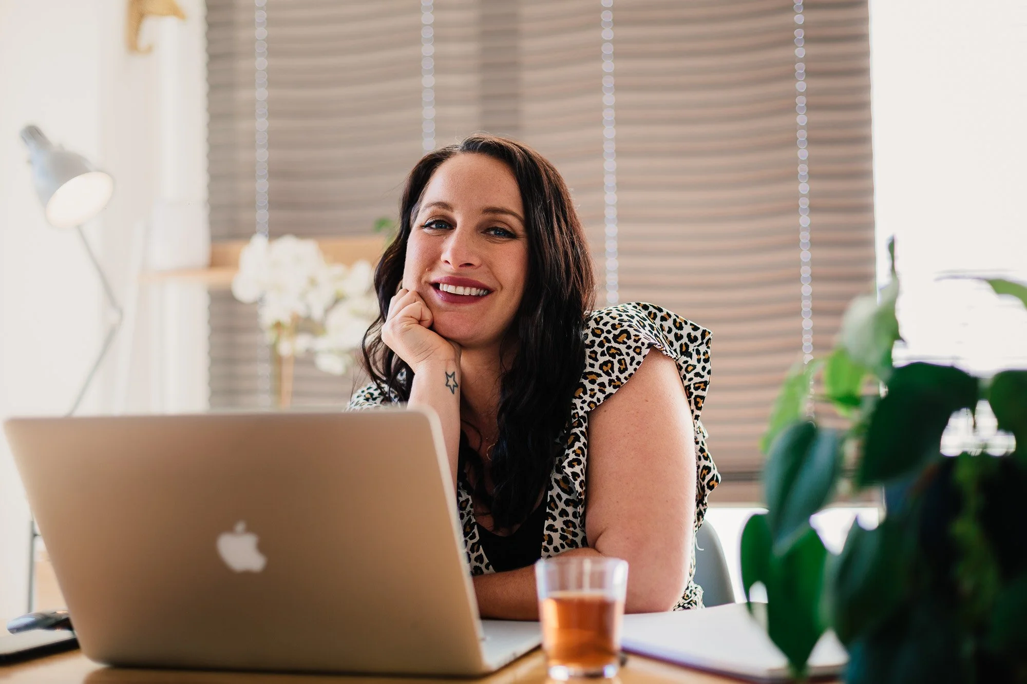 A woman sitting at a desk with an open MacBook laptop, smiling and resting her chin on her hand, in a bright office with a window, blinds, a potted plant, and a glass of tea.