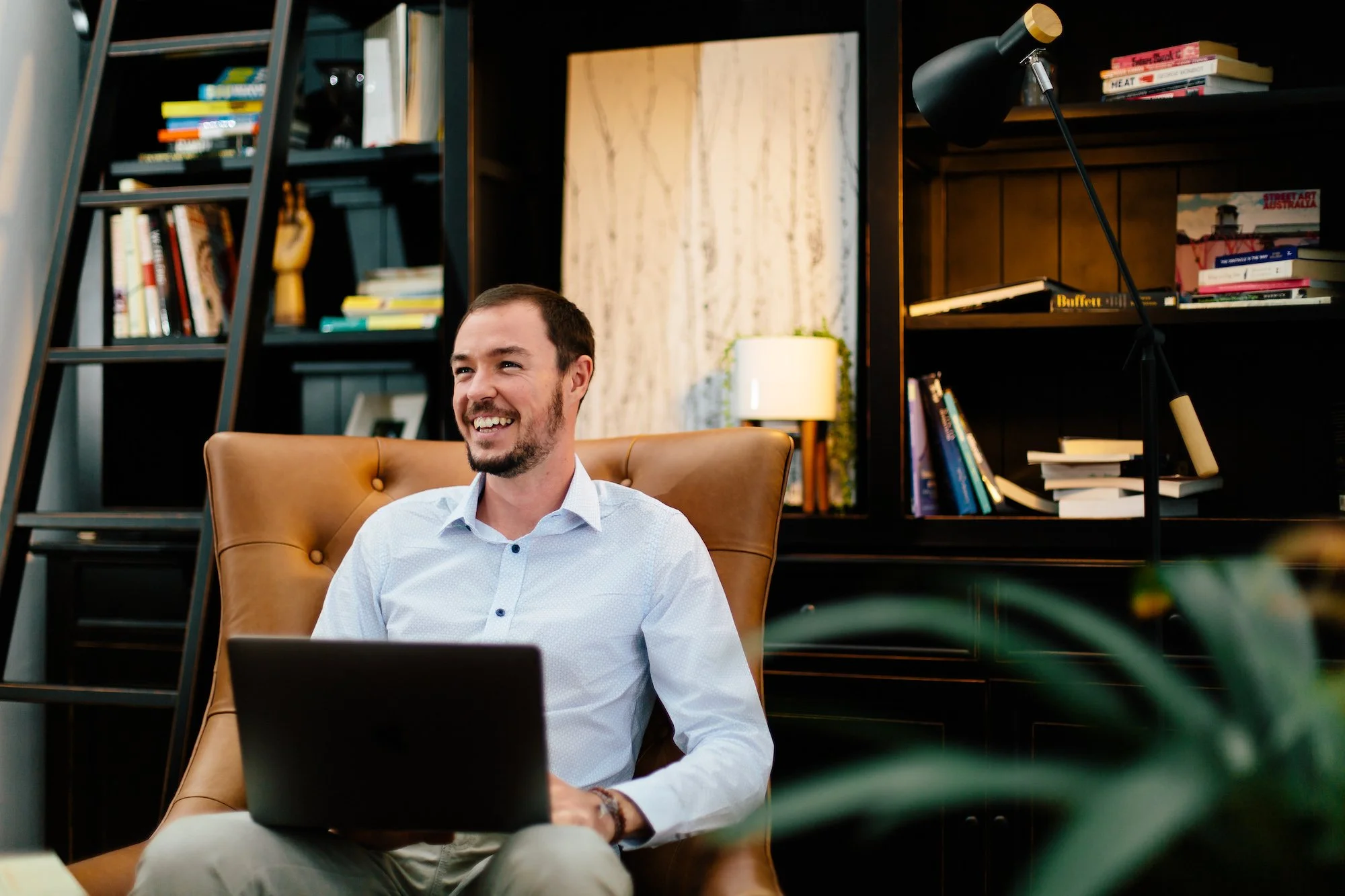 A man smiling while sitting in a leather chair with a laptop on his lap in a cozy study or library with dark bookshelves, books, and a lamp in the background.