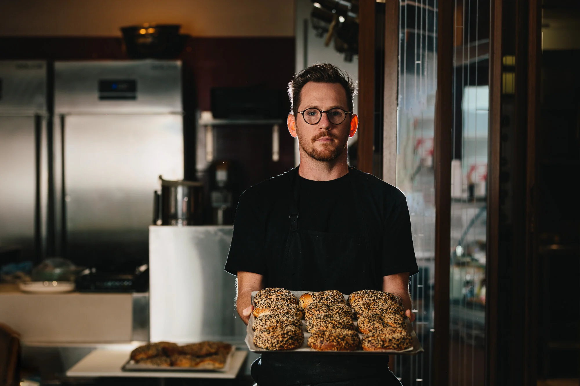 A man wearing black clothes and glasses holding a tray of sesame seed-coated bagels in a bakery or kitchen setting.