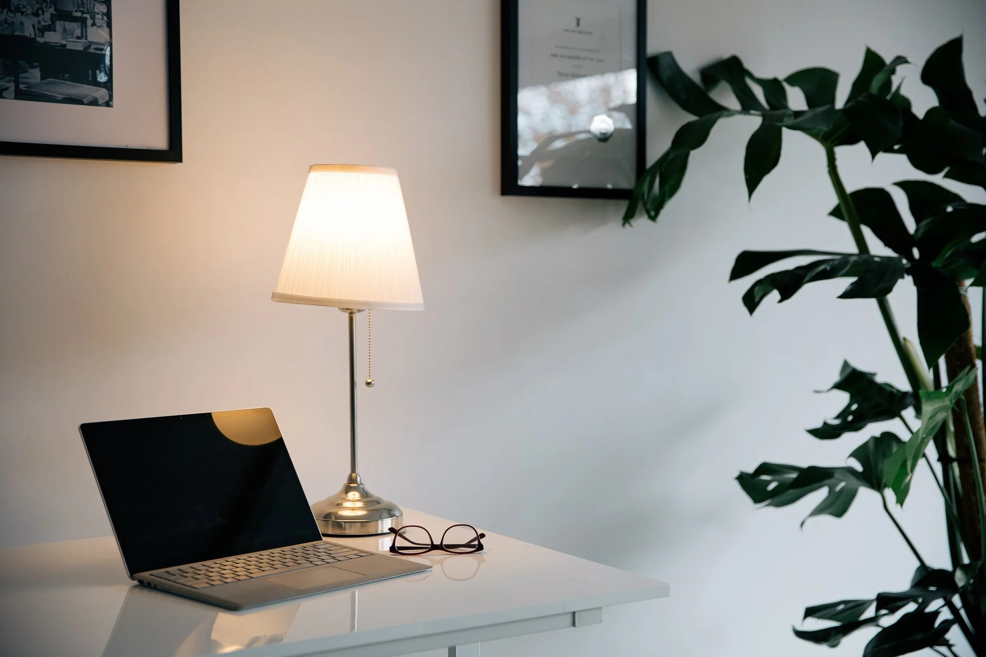 A white desk with a laptop, a pair of glasses, and a lit table lamp. There are framed pictures on the wall and a large potted plant on the right.