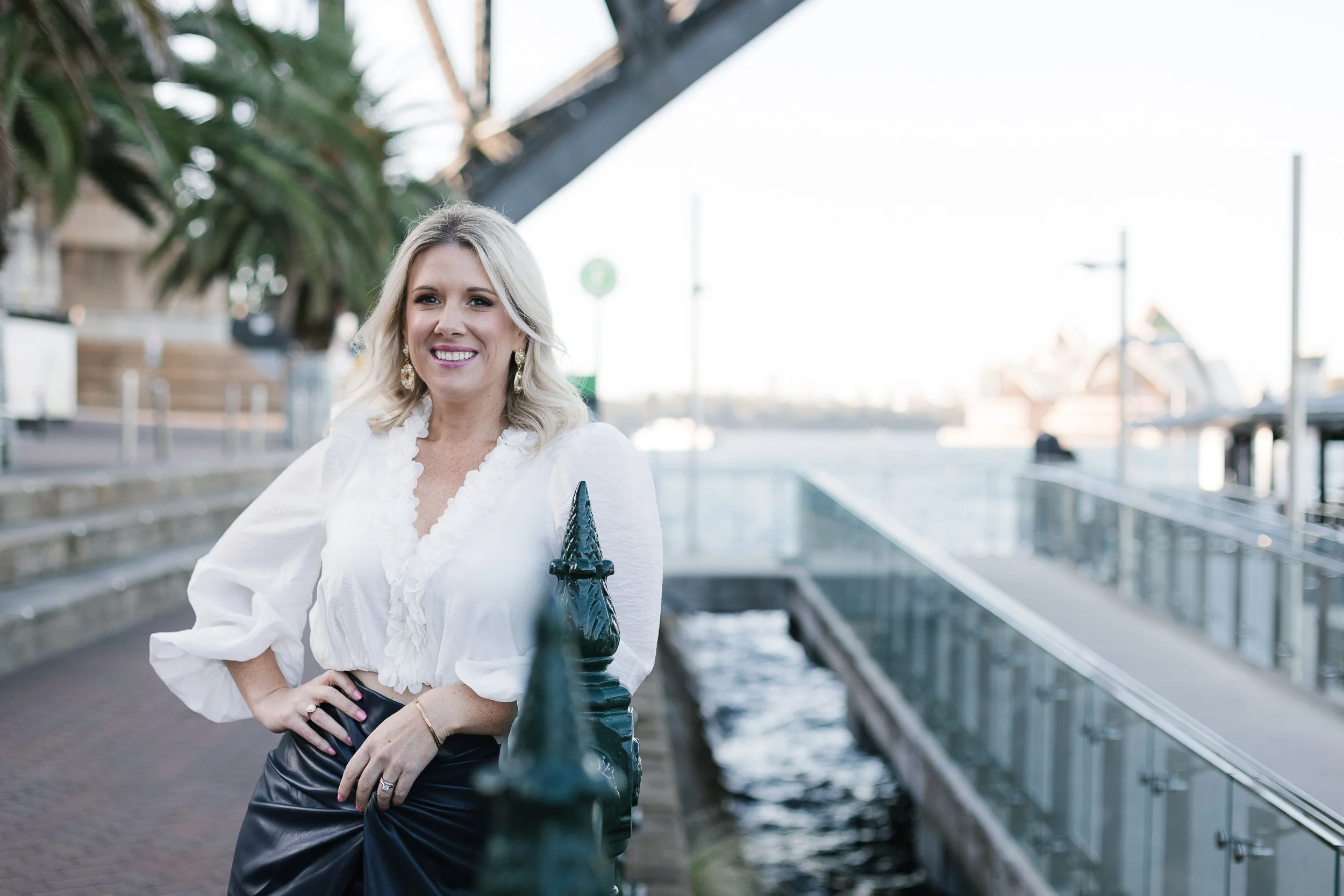 A woman with blonde, wavy hair wearing a white blouse and earrings, standing outdoors by a railing near water with boats and palm trees in the background.