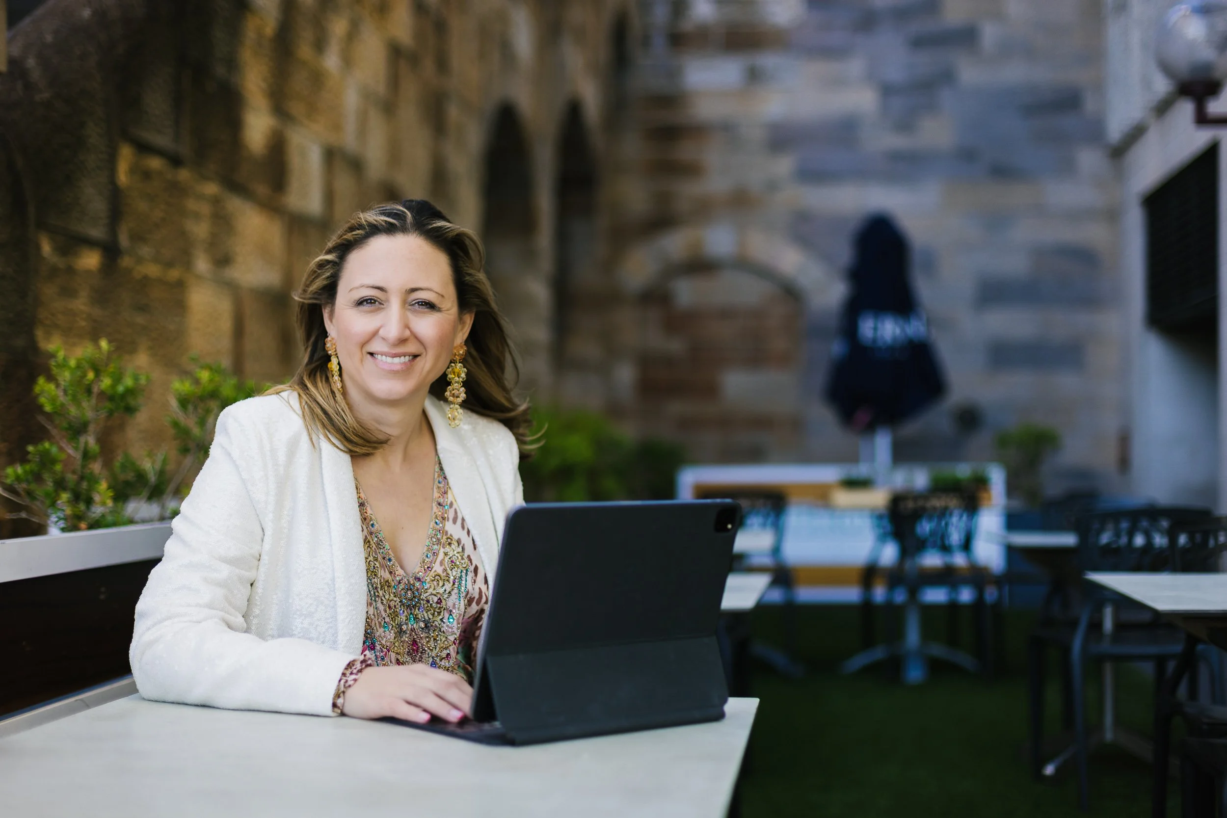 A woman with shoulder-length brown hair, wearing a white blazer and large gold earrings, sitting at a table outdoors, smiling at the camera with a tablet in front of her.