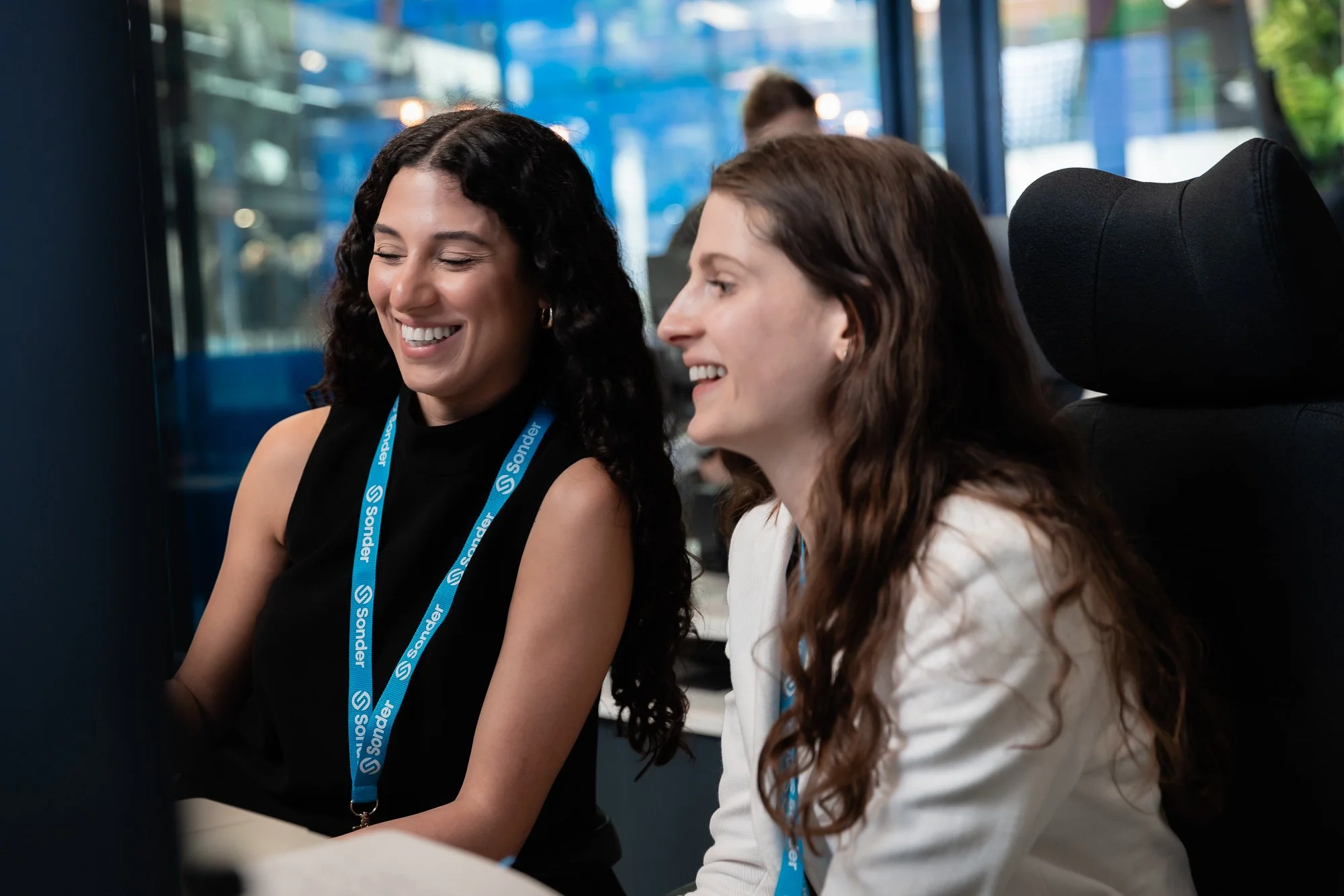 Two women smiling and chatting at a business event, wearing lanyards with the name 'Sonder', in a modern office setting.