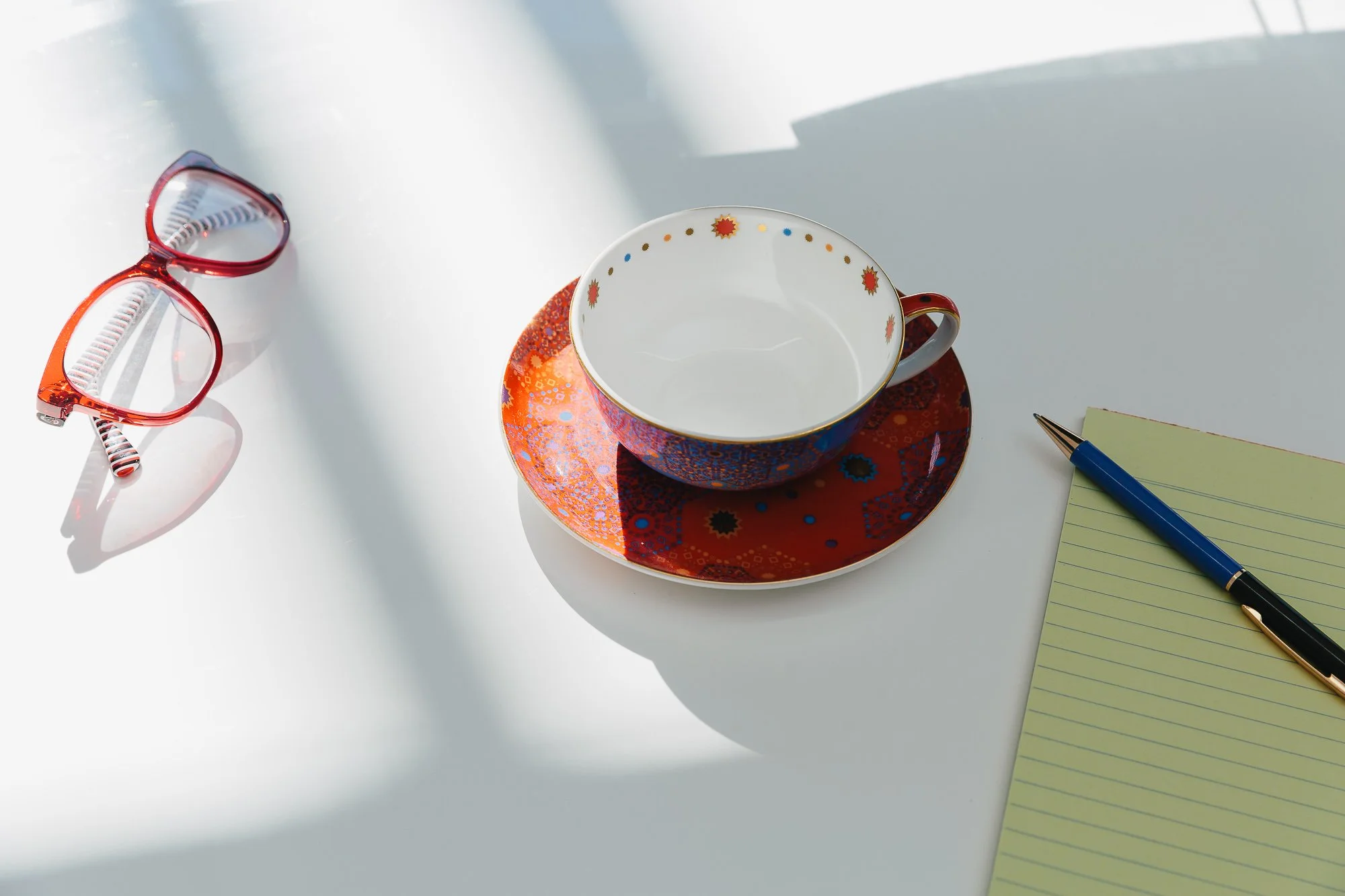 Red and blue patterned teacup on matching saucer, yellow notepad with black and gold pen, red glasses on white surface.