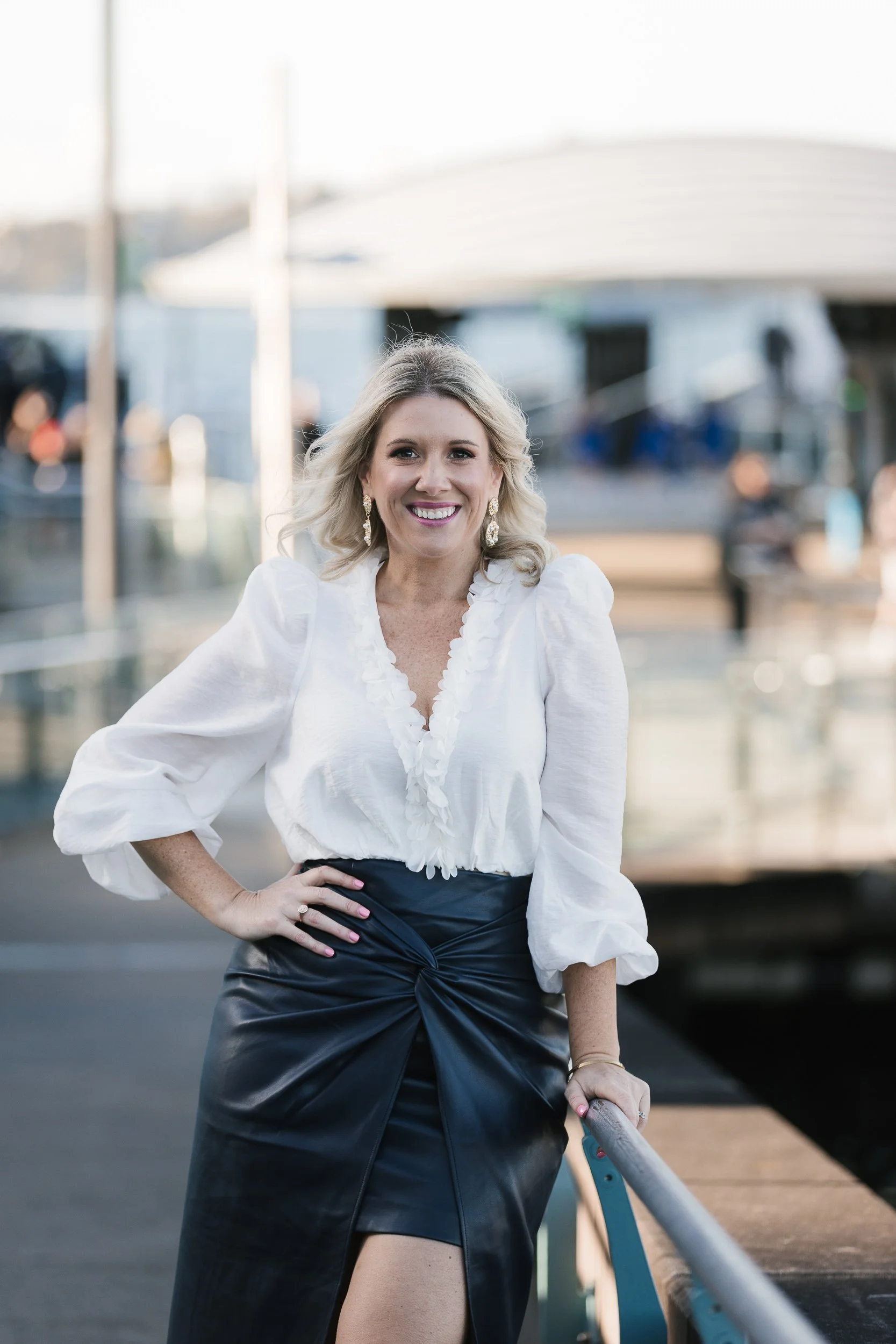 A smiling woman in a white blouse and black leather skirt, standing outdoors near a marina with boats in the background.