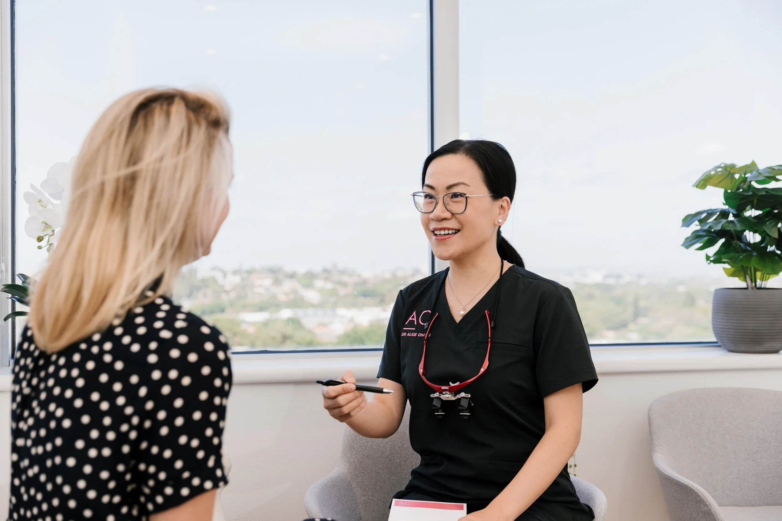 A healthcare professional, wearing black scrubs and glasses, smiling while speaking to a patient with blonde hair in a polka-dot blouse, inside an office with a large window and a potted plant in the background.