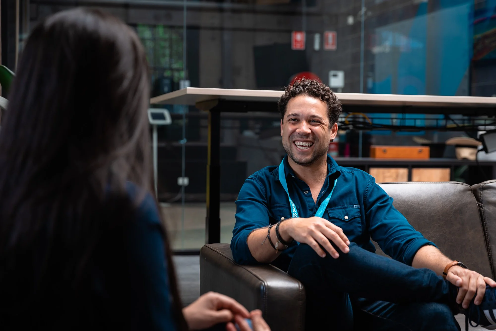 Man with curly hair and earrings smiling and talking to a woman with dark hair in a casual indoor setting.
