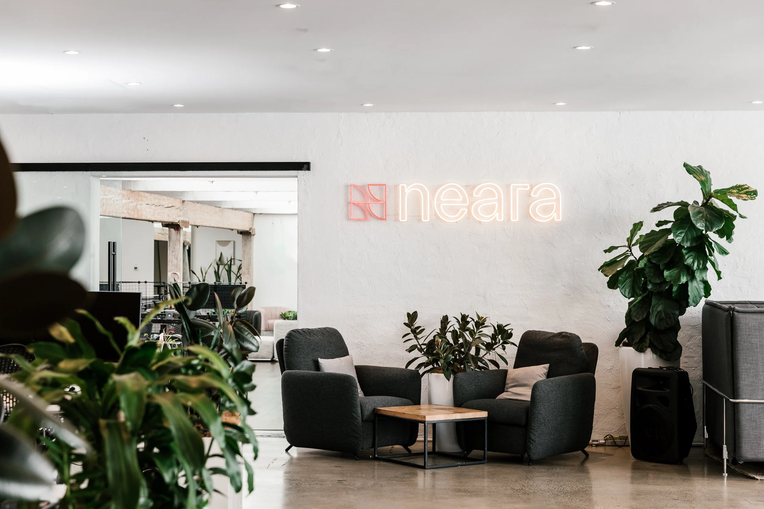 Modern interior with black armchairs, potted plants, a small table, and a neon sign that says 'neara' on a white wall.