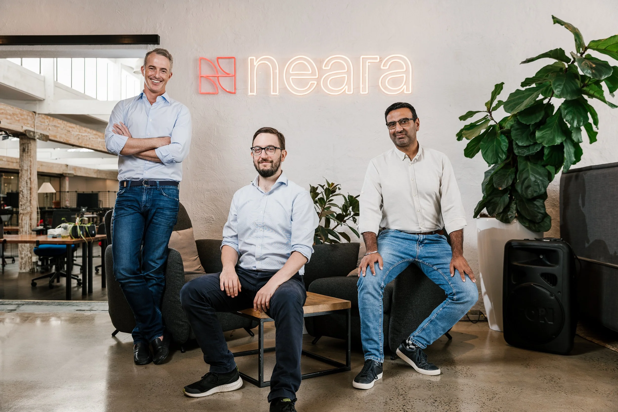 Four men in casual business attire posing in an office lounge area with a neon sign reading 'neara' on the wall behind them. The space has modern decor, plants, and a speaker on the floor.