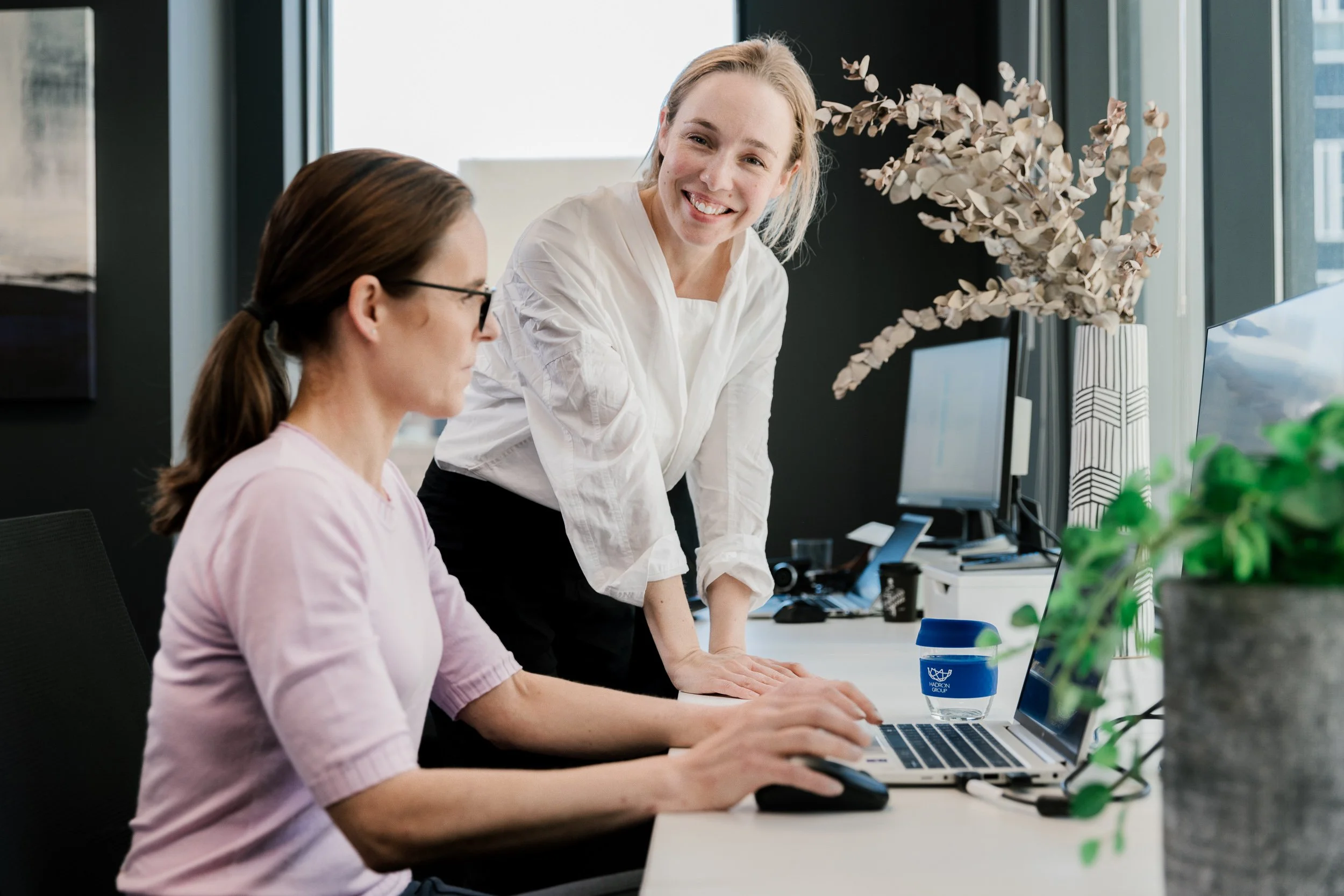 Two women working in an office, one sitting and using a laptop, the other standing and smiling.