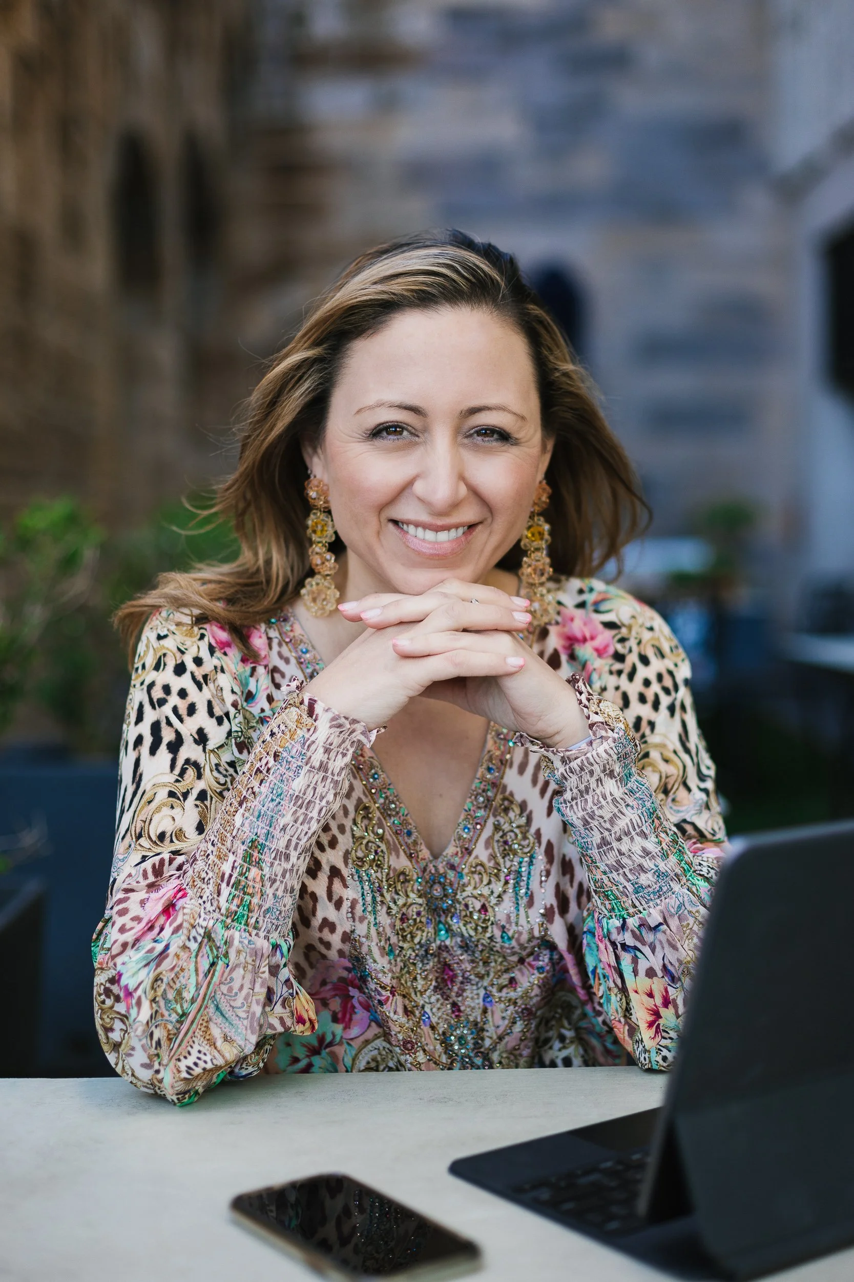 A smiling woman with light brown hair, wearing large earrings and a colorful, patterned top, sits at a table outdoors with a laptop, smartphone, and blurred background.