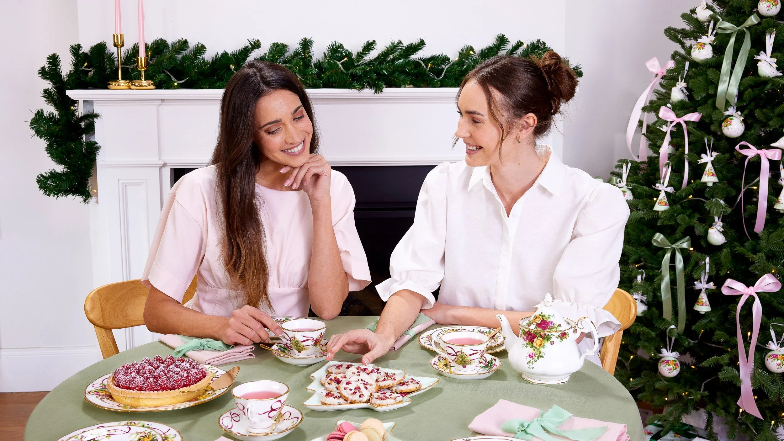 Royal Albert. Two women sitting at a table setting drinking tea with a Christmas tree and fireplace behind them.