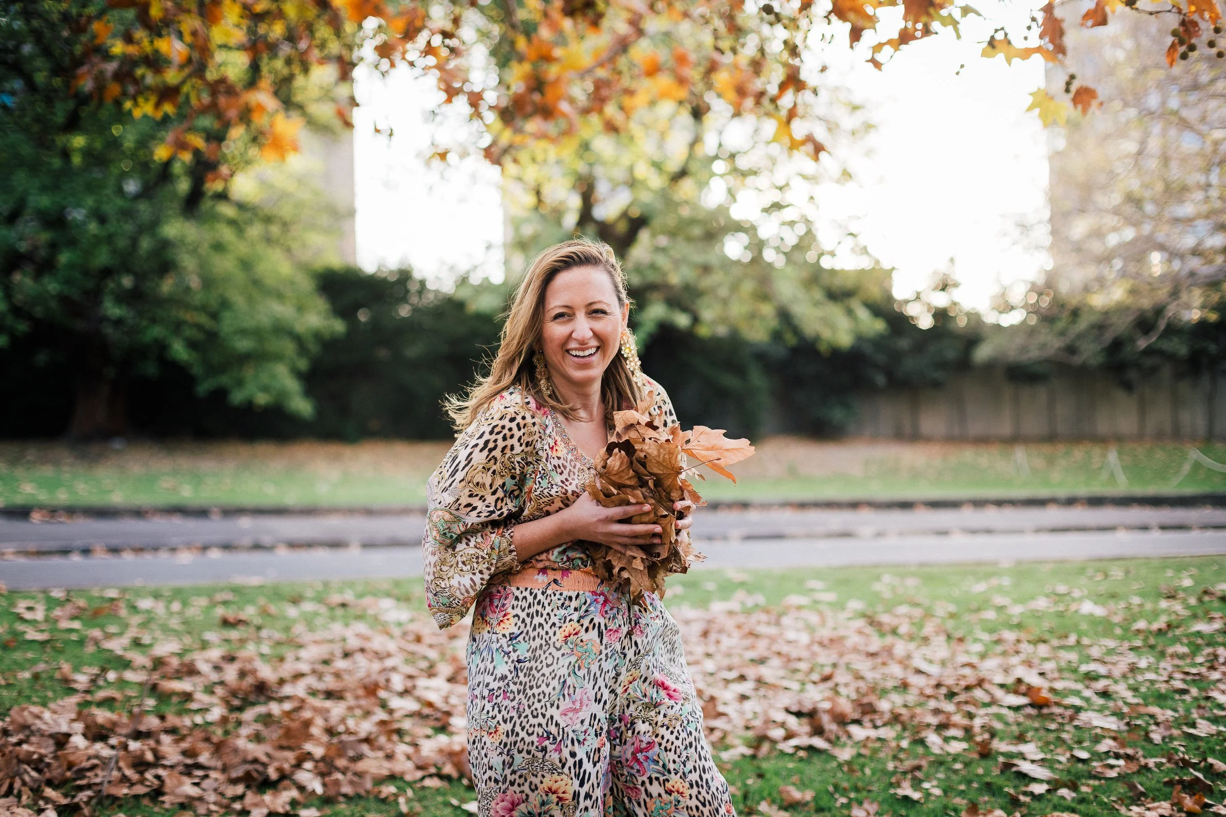 A woman standing outdoors on a fall day, holding a bunch of brown leaves, smiling, with trees and fallen leaves in the background.