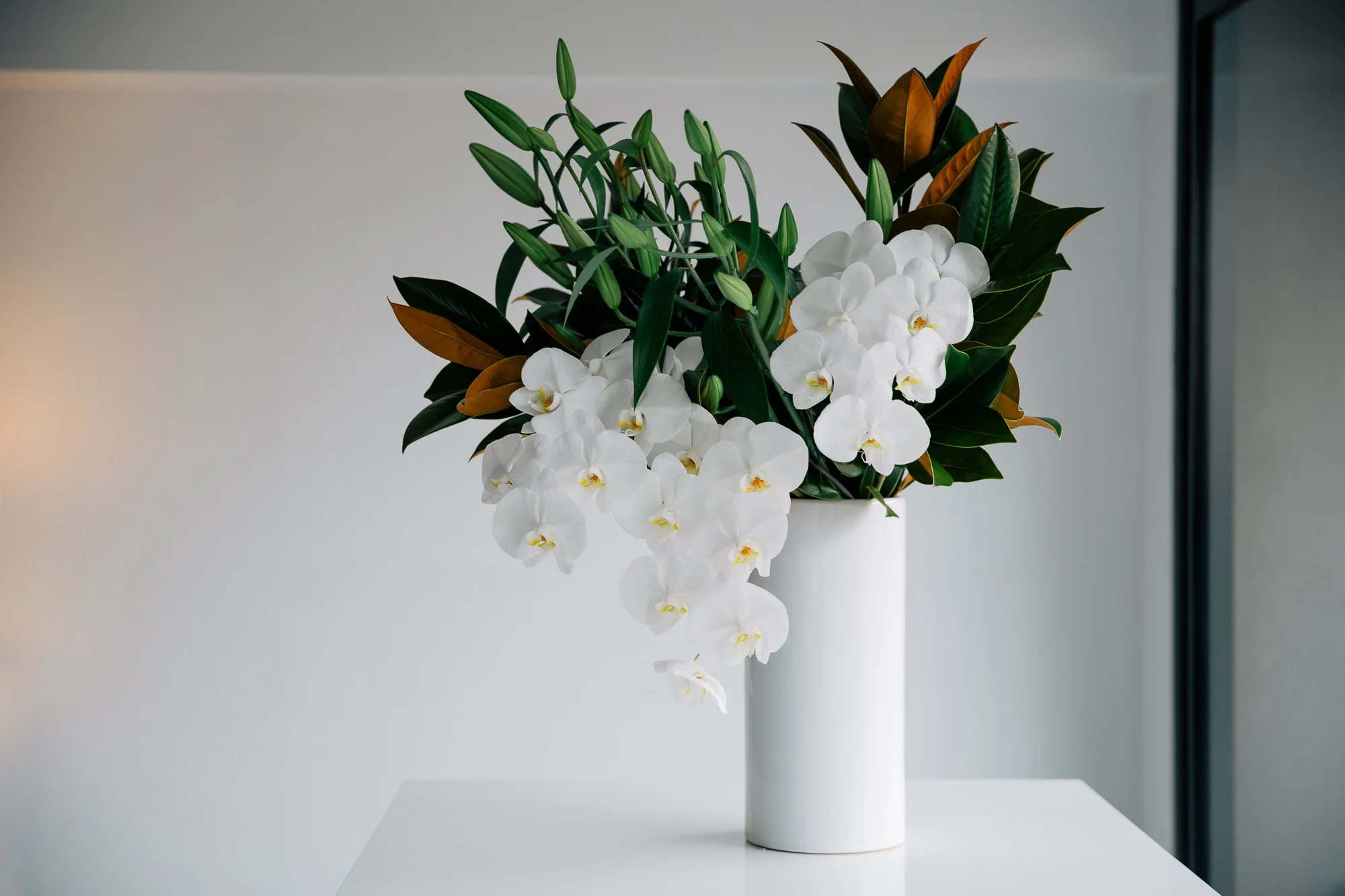A white ceramic vase with a bouquet of white orchids, green leaves, and brown-tipped leaves on a white surface against a plain light-colored background.
