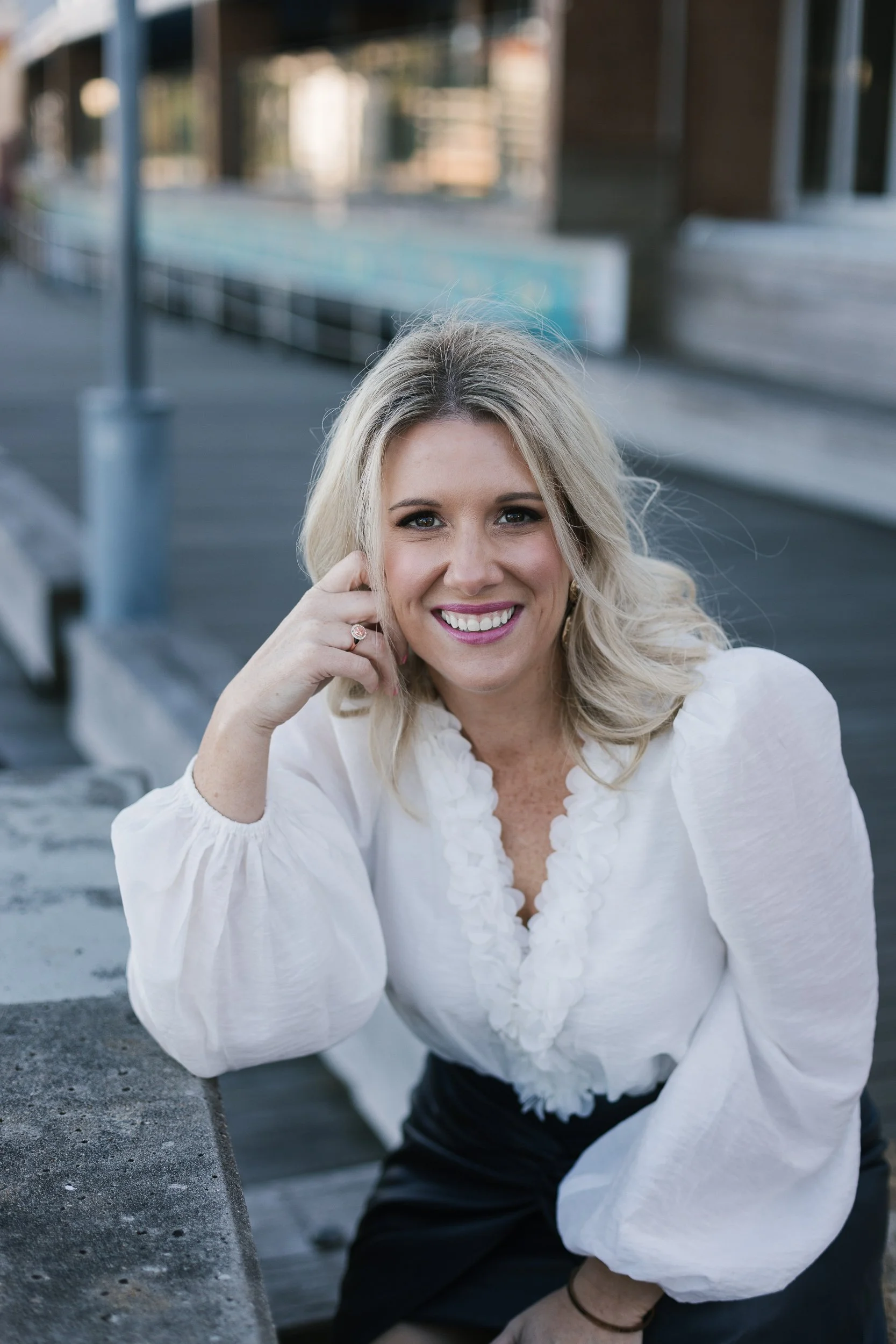 A woman with blonde hair smiling outdoors, wearing a white blouse and black skirt.