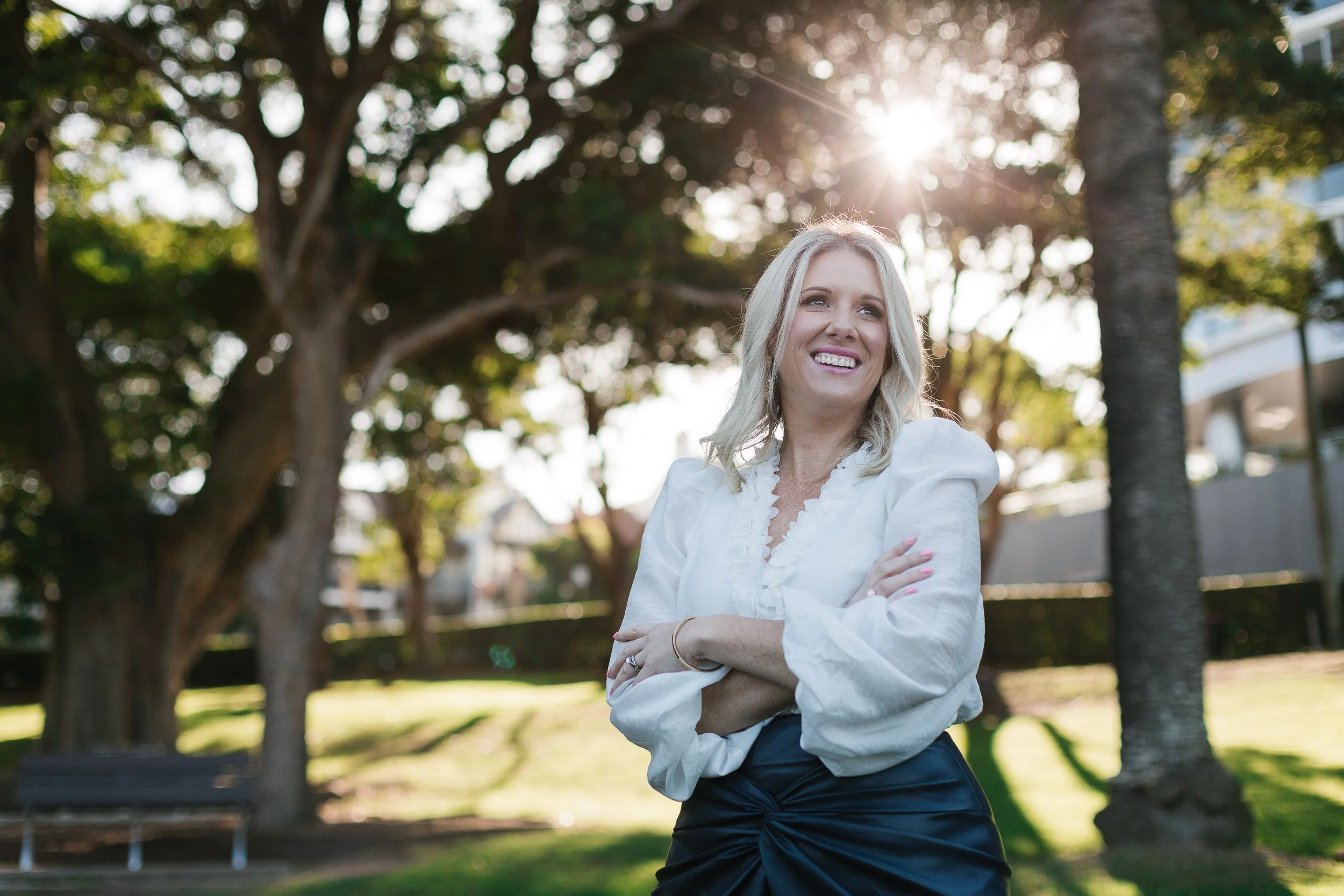 A smiling blonde woman in a white blouse and dark skirt standing outdoors in a park with trees and sunlight in the background.
