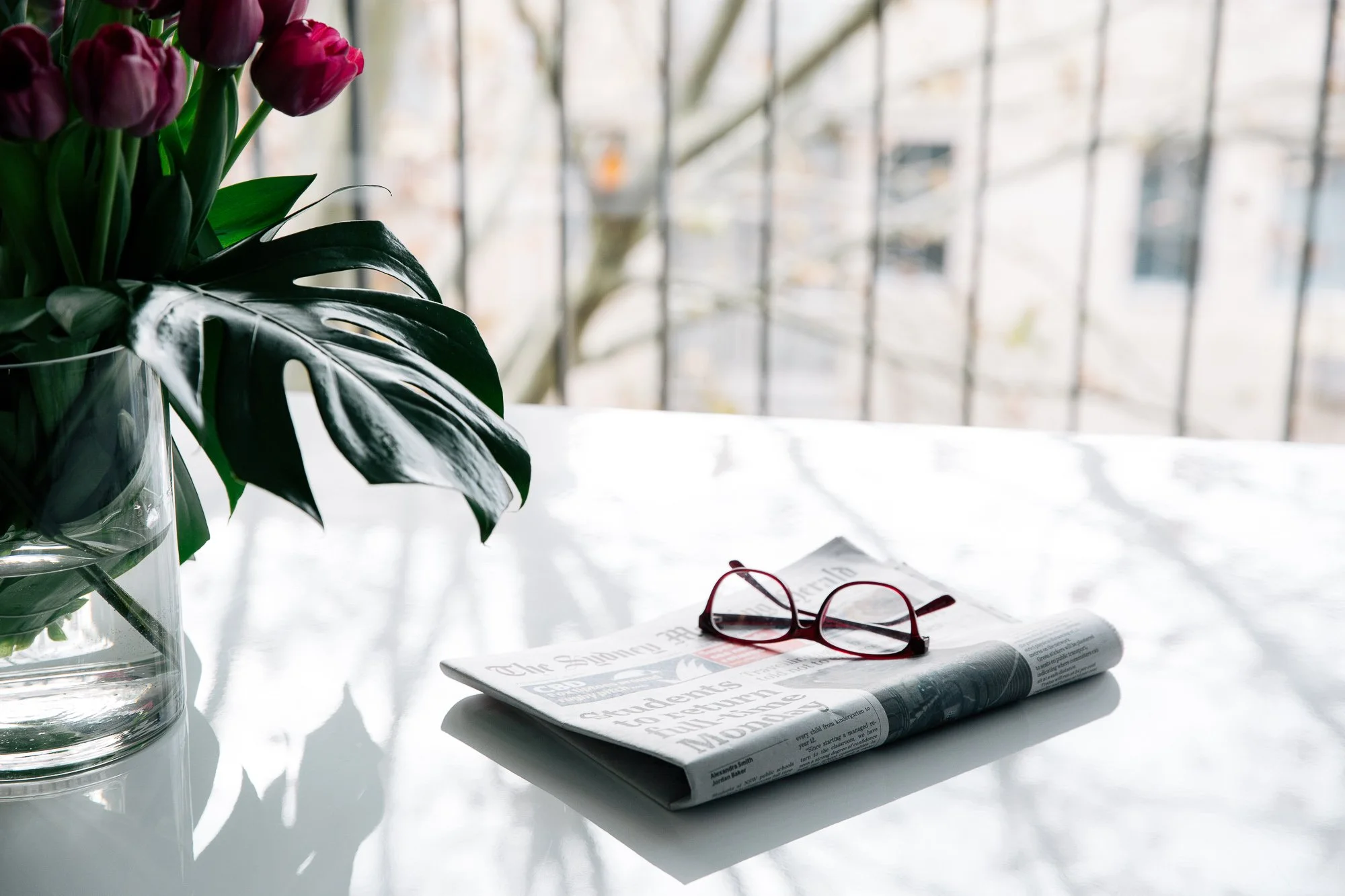 A glass vase with purple tulips and a large dark green Monstera leaf on a reflective white table. Next to it, a folded newspaper with red glasses resting on top. In the background, a large window with a grid pattern shows an outdoor scene with trees 