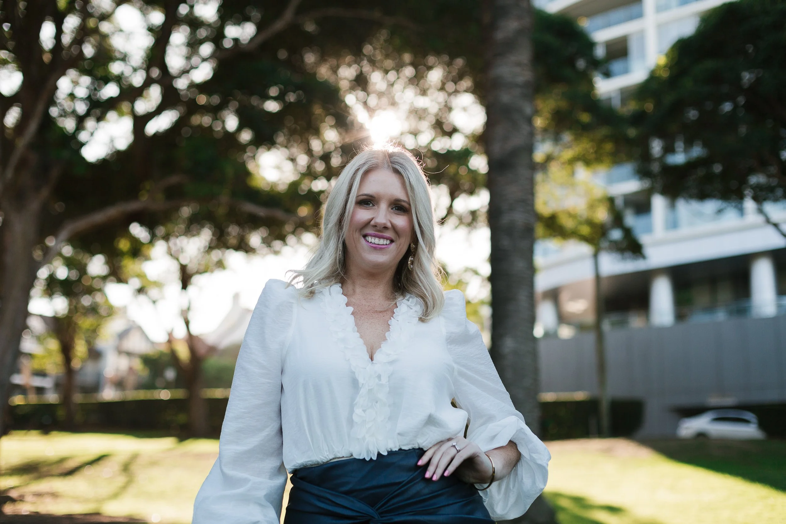 A woman with blonde hair smiling outdoors in front of trees and a modern building, sunlight shining behind her.