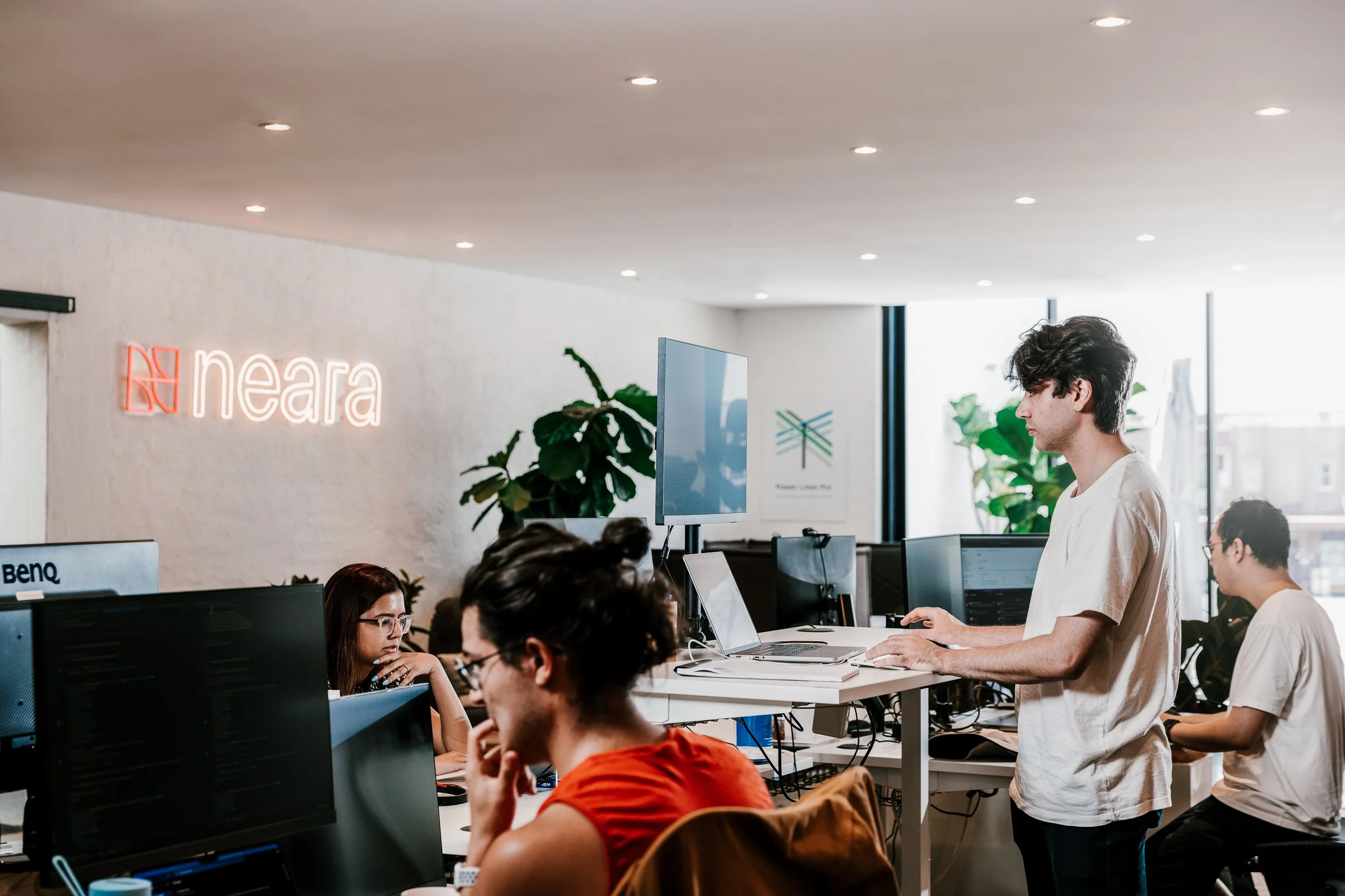 Office scene with multiple people working at computers, a standing person at a desk, large windows, plants, neon 'neara' logo on the wall.