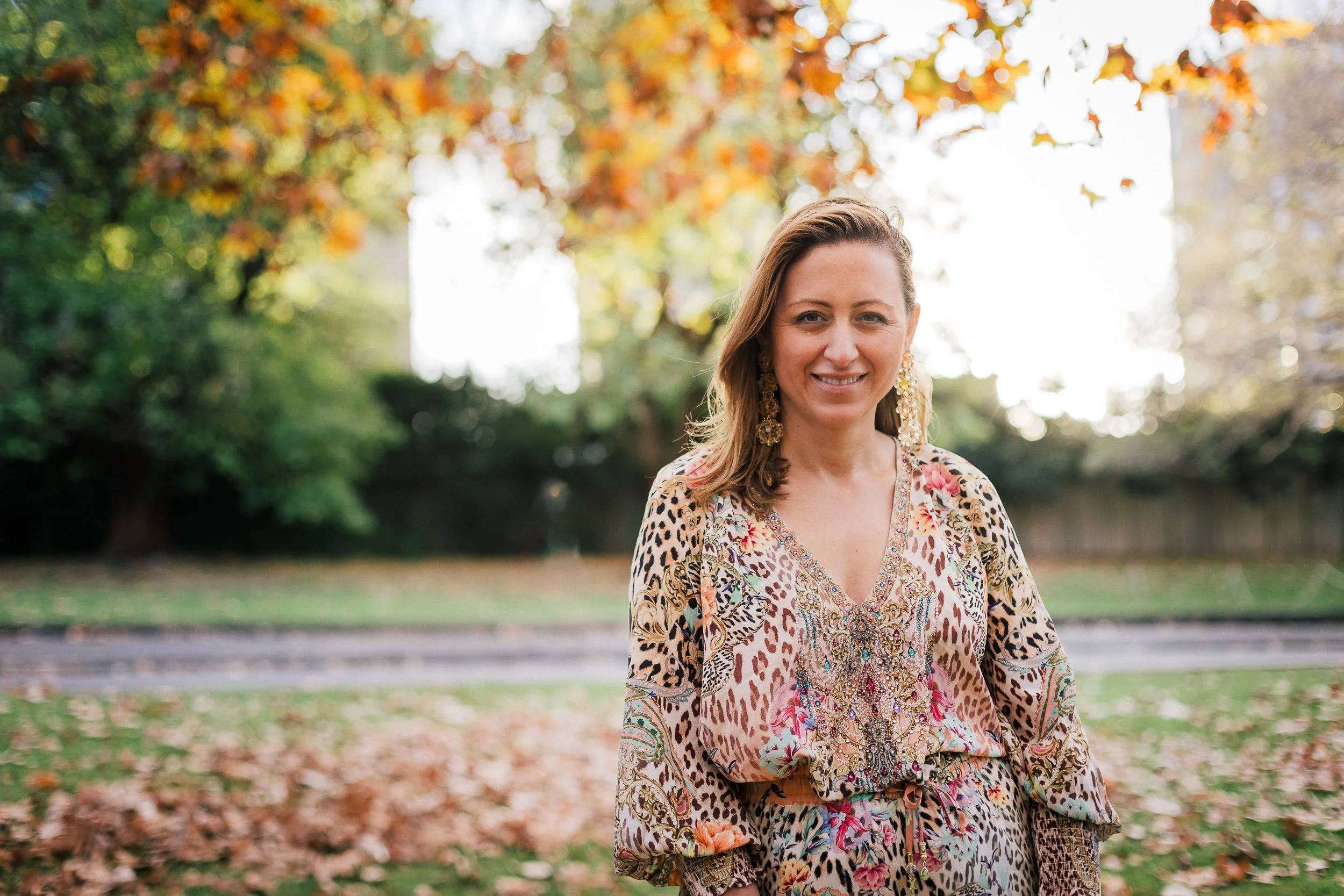 A woman with shoulder-length hair, wearing a colorful patterned dress and large earrings, standing outdoors in a park with autumn leaves on the ground and trees in the background, smiling at the camera.