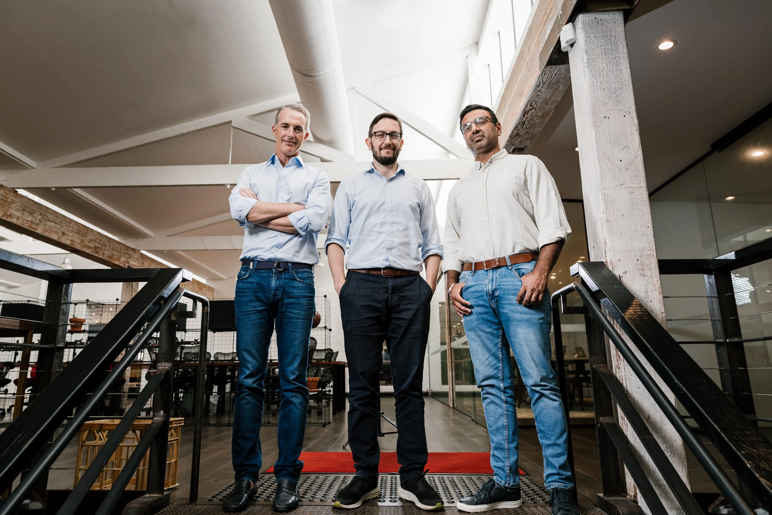 Three men standing in a modern office space on a red carpet, with arms crossed or in pockets, looking at the camera.