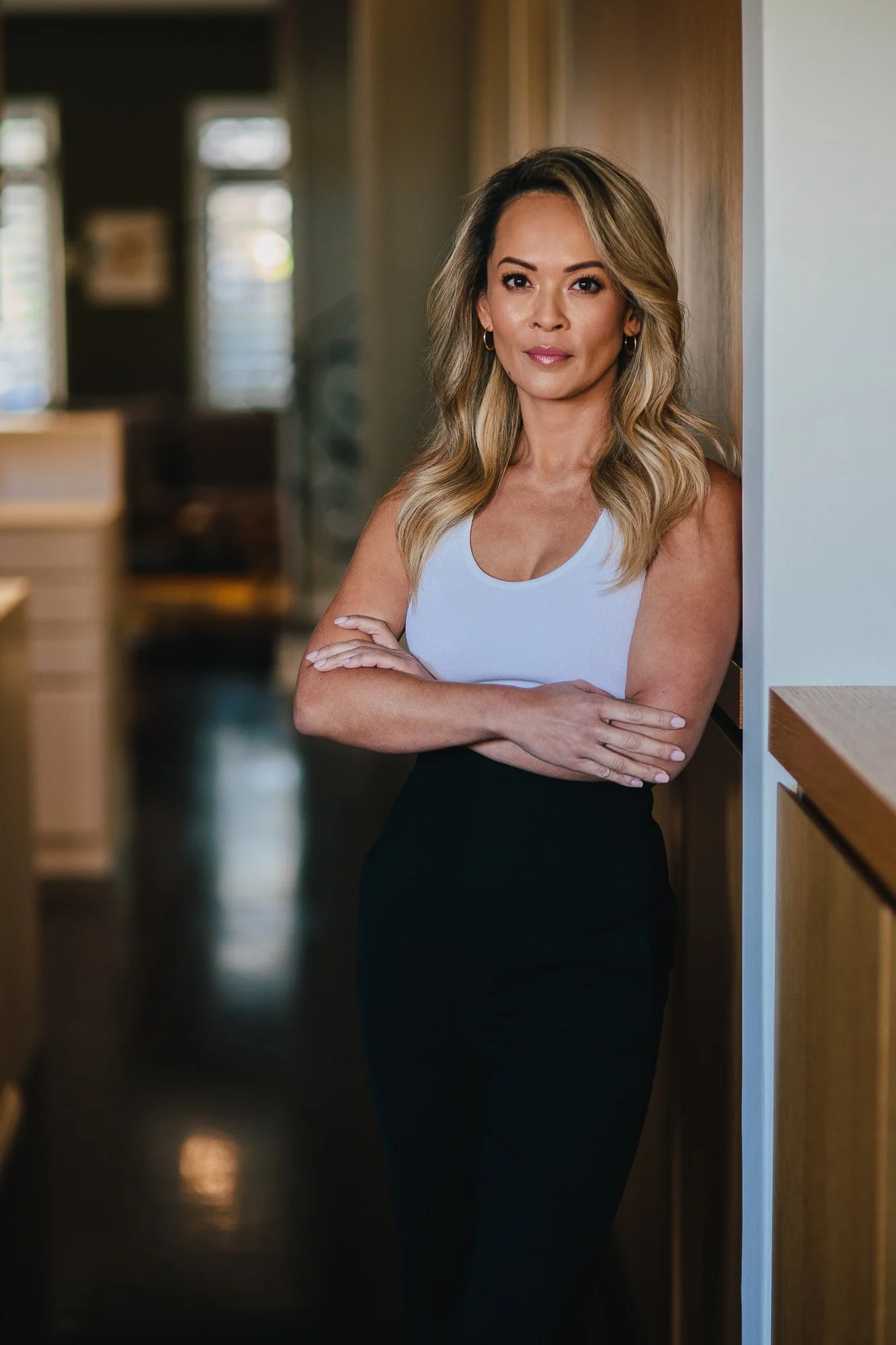A woman with blonde wavy hair dressed in a white sleeveless top and black high-waisted pants standing indoors with arms crossed, looking at the camera.