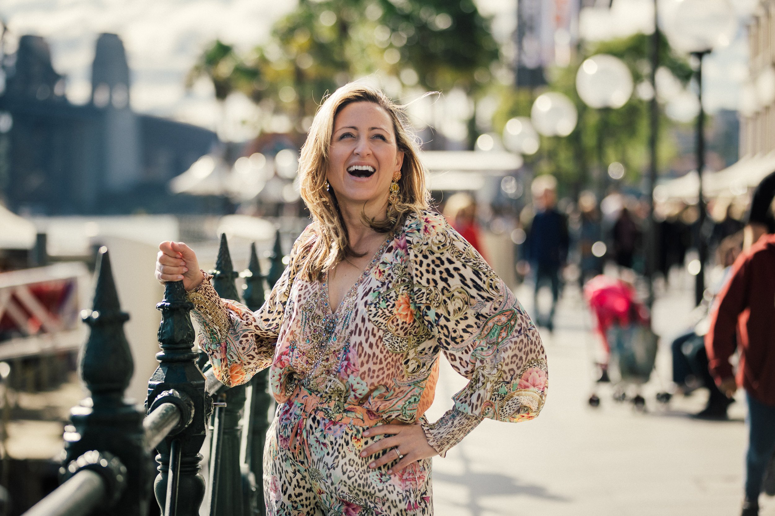 A woman with blonde hair wearing a colorful, patterned dress, smiling and holding onto a fence in an outdoor urban setting with blurred people and trees in the background.