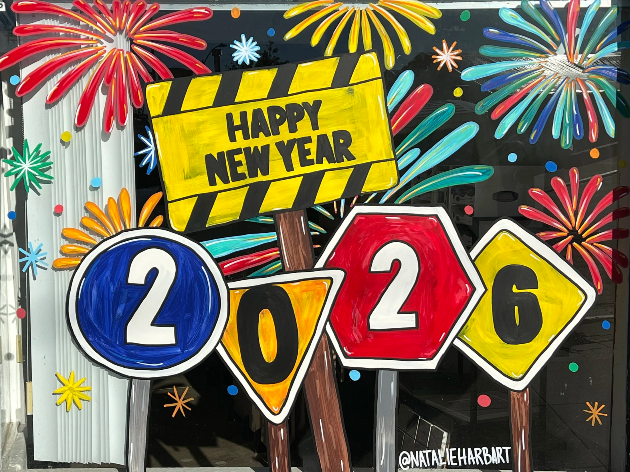 Colorful New Year's Eve window display with fireworks, a yellow sign saying 'HAPPY NEW YEAR,' and numbers 2026 with varied shapes and colors, including blue, red, and yellow. Created by @NATALLIEHARBART.