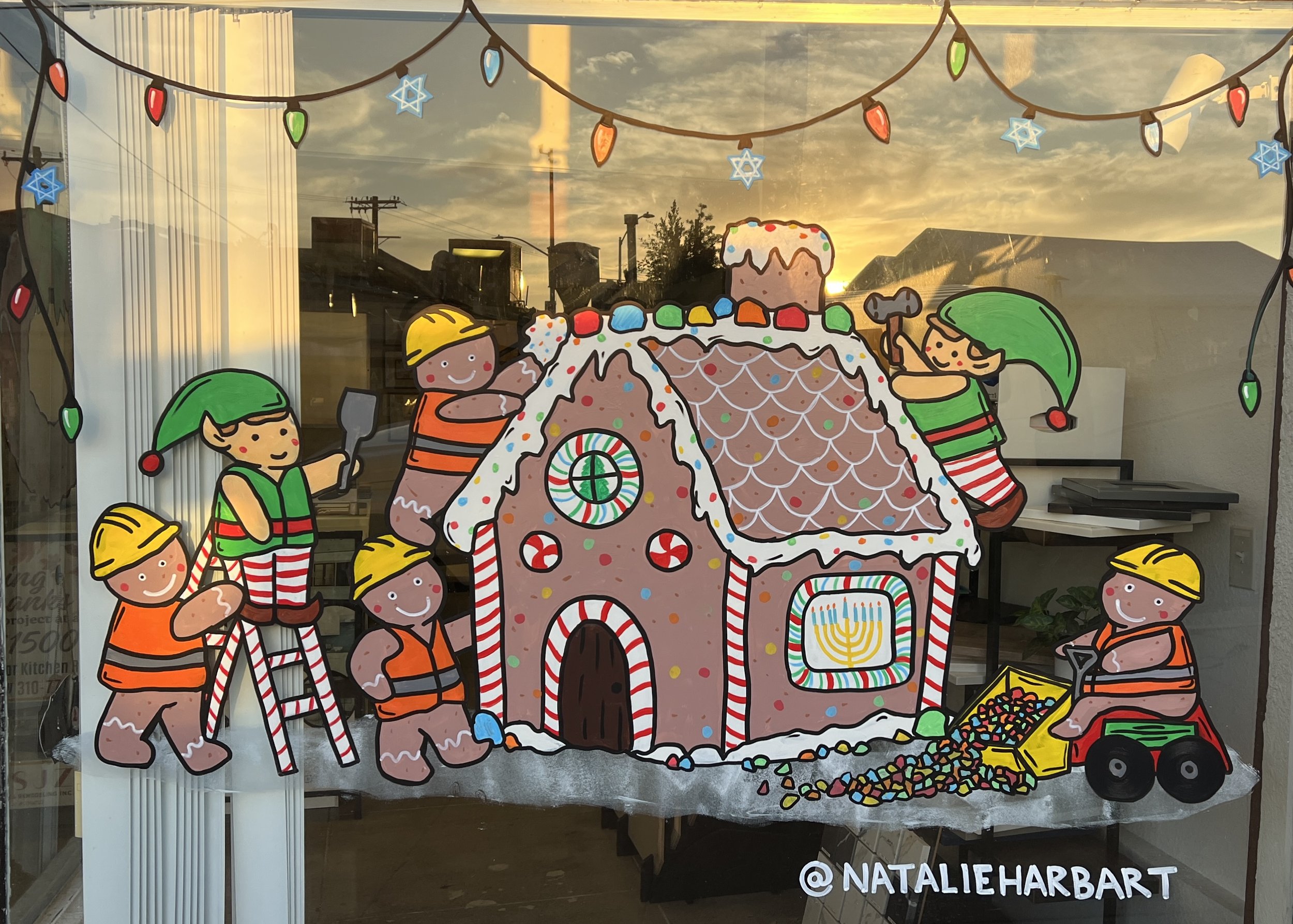 Colorful holiday window display featuring a gingerbread house with candy decorations, surrounded by cartoon elves and children, with string lights and a sunset background.